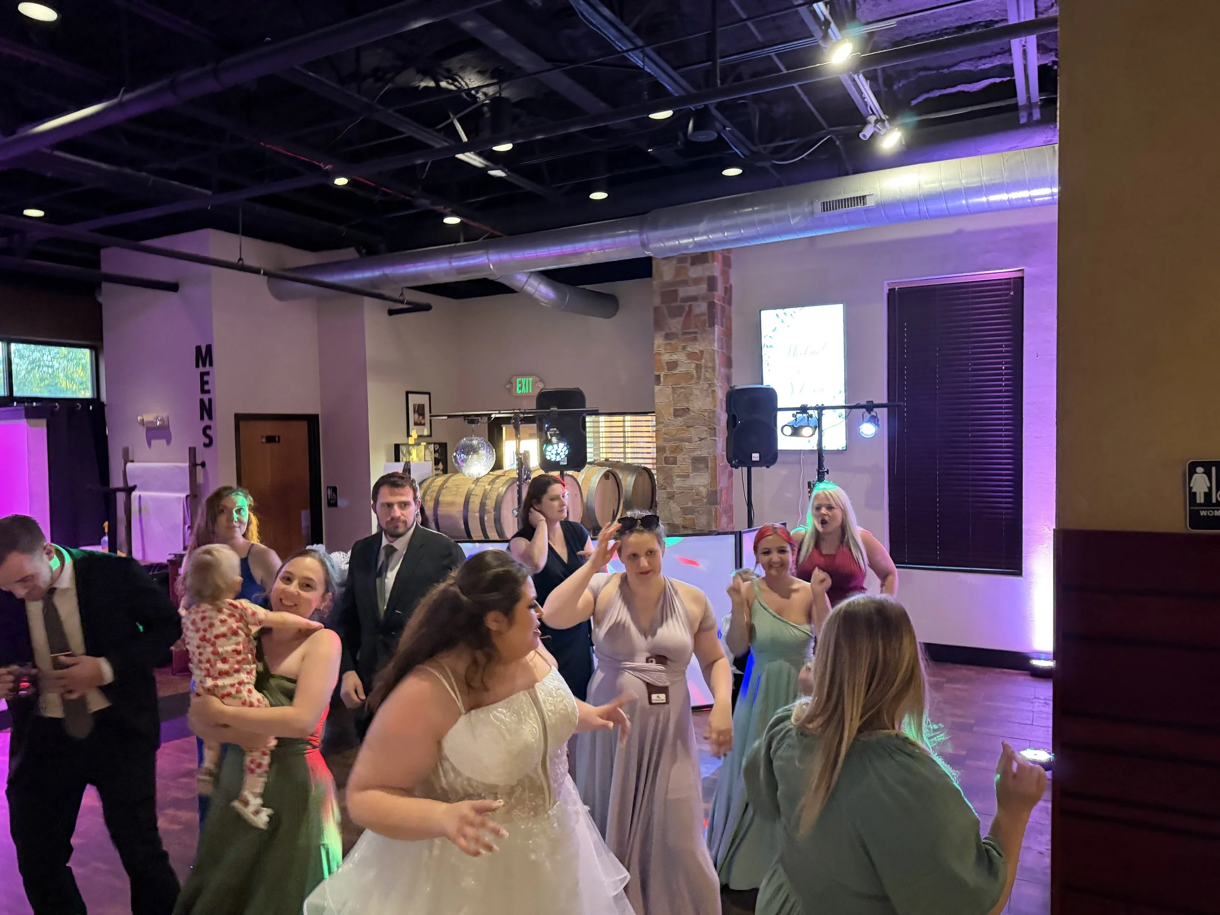 People dancing and celebrating in a dimly lit indoor venue decorated with wine barrels and colorful lights.