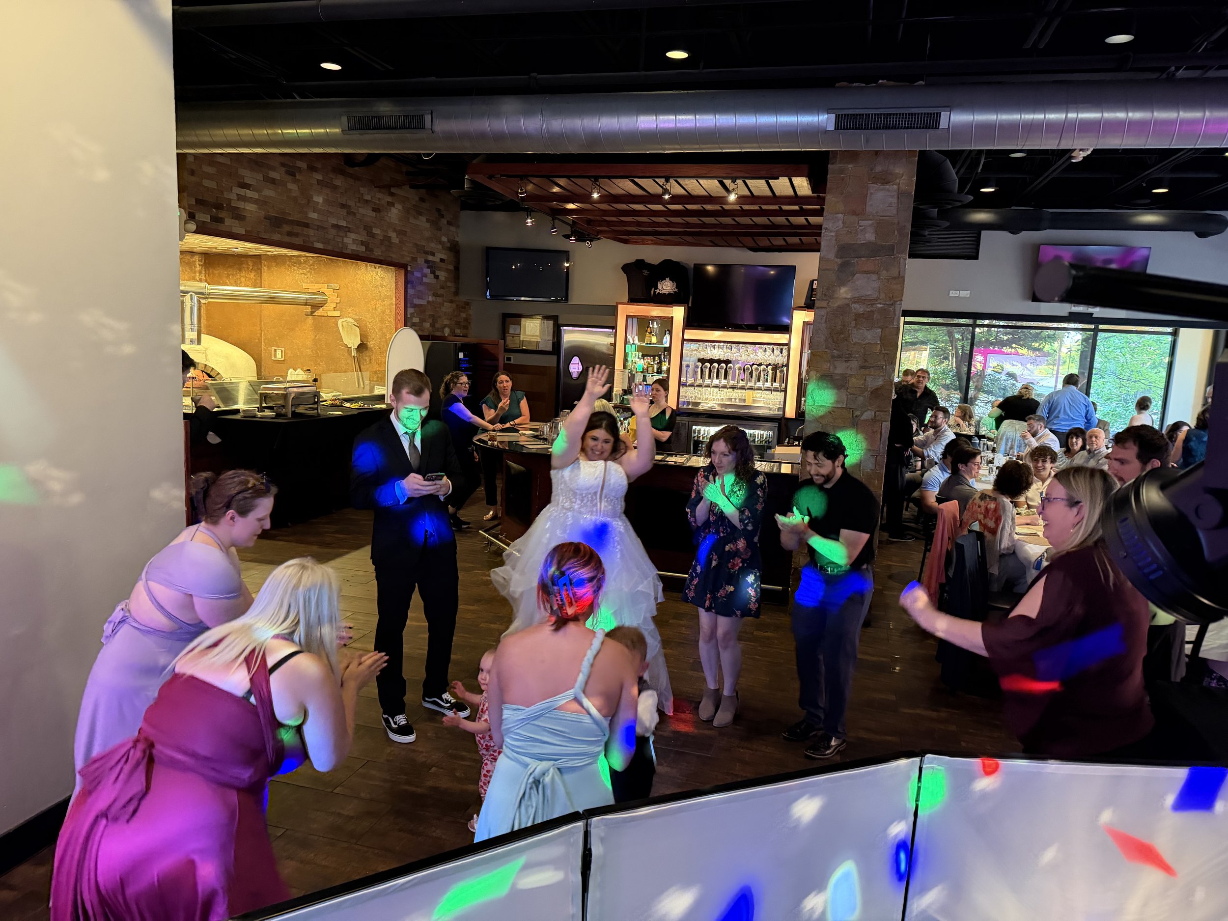Guests at a celebration or wedding reception dancing and socializing inside a restaurant or event space with brick walls, large windows, and a bar area.