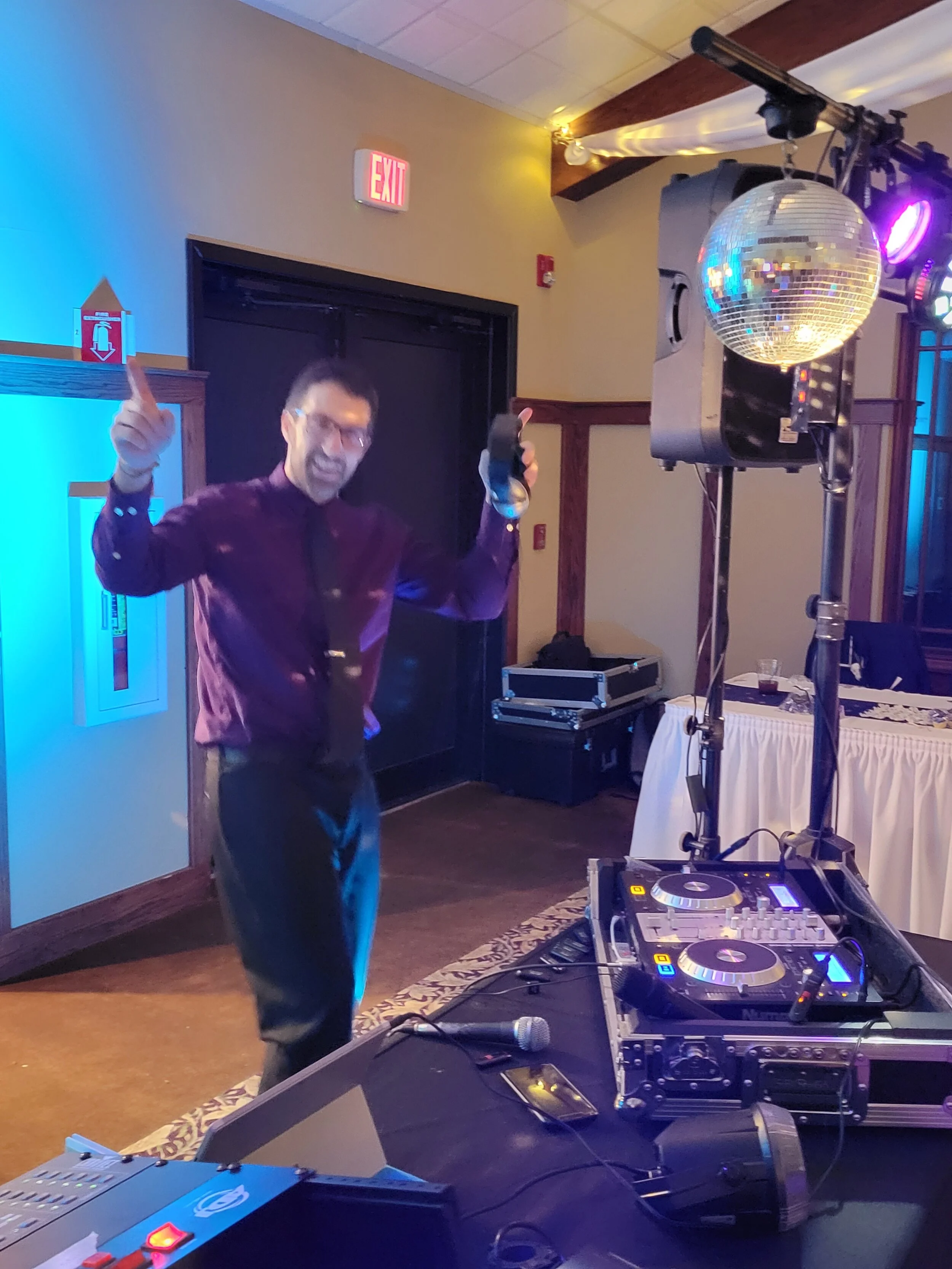 DJ setup with turntables and gear, person dancing nearby, disco ball and stage lighting, indoor event.