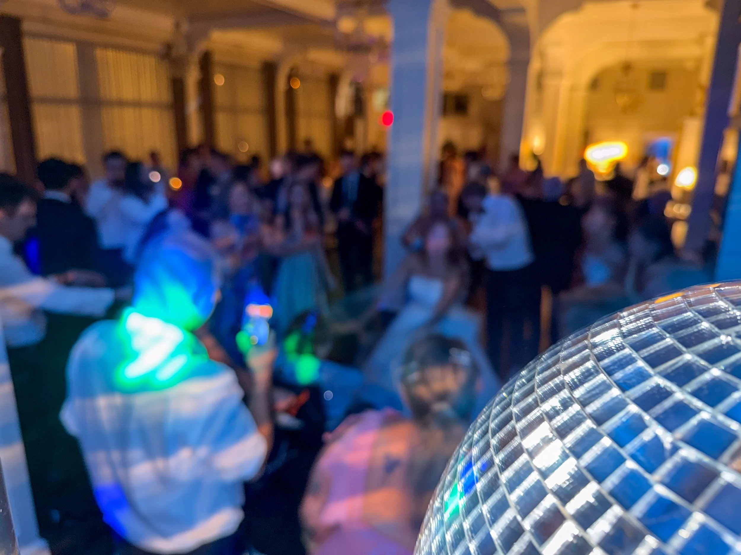 A large group of people, some dressed in formal attire and others casual, are gathered indoors. The lighting is dim, with some colorful lights creating a festive atmosphere. The setting appears to be a social event or party in a spacious hall with hi