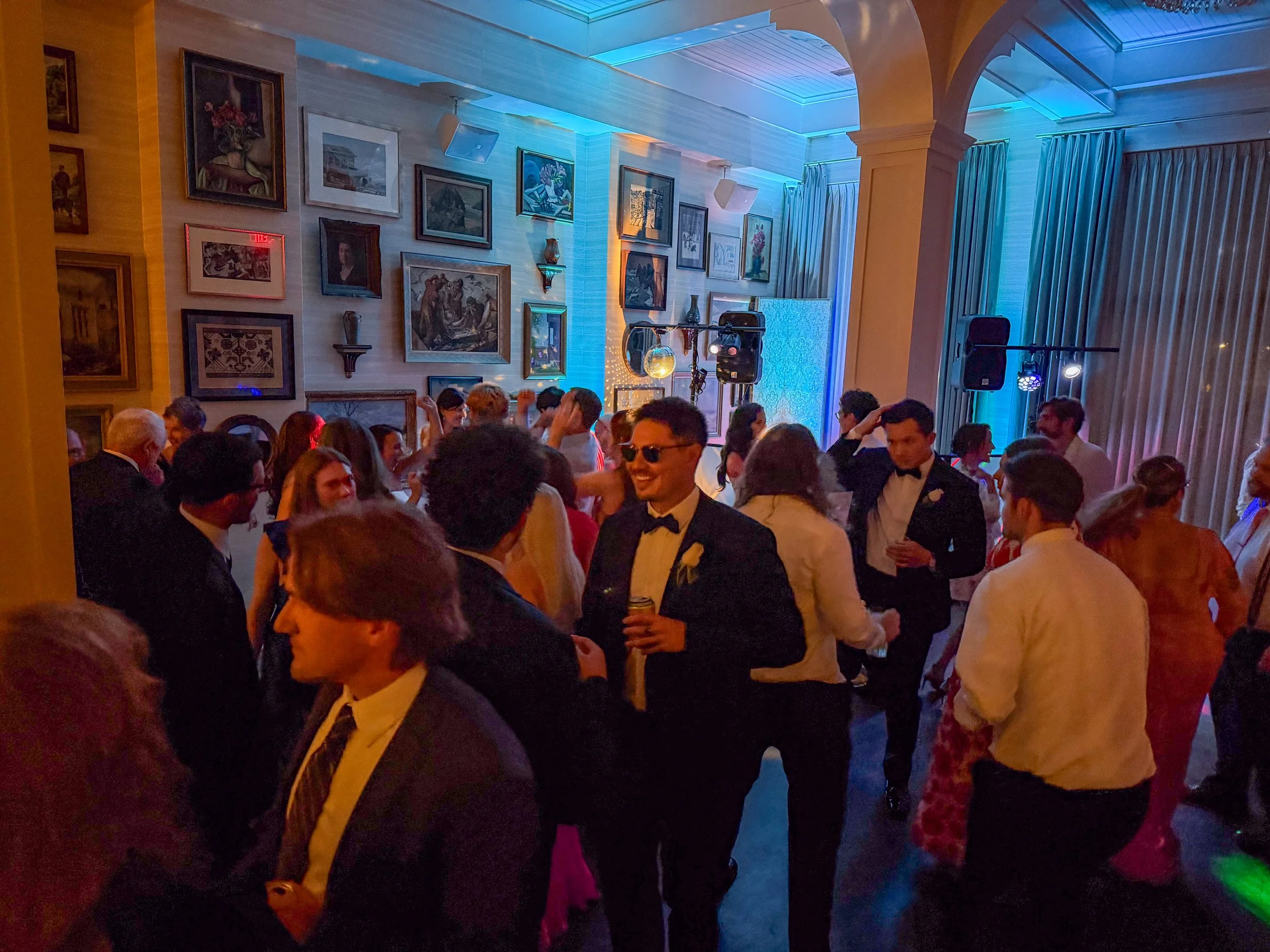 A group of people dressed in formal attire, such as tuxedos and dresses, dancing and celebrating at an indoor event with many paintings on the wall and blue lighting.