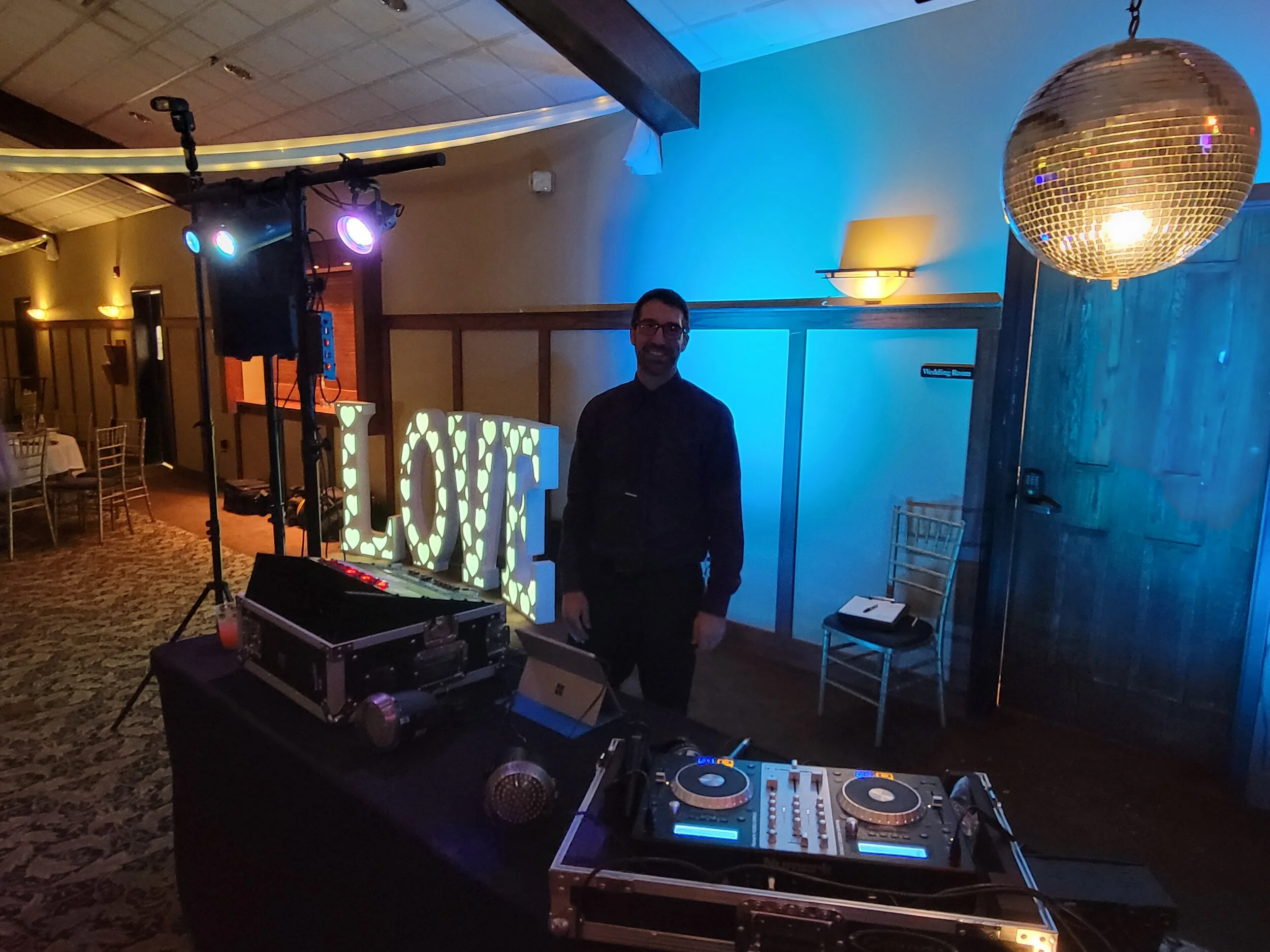 DJ setup at an event with lighting equipment, DJ console, and a large "LOVE" sign. Disco ball hangs above.