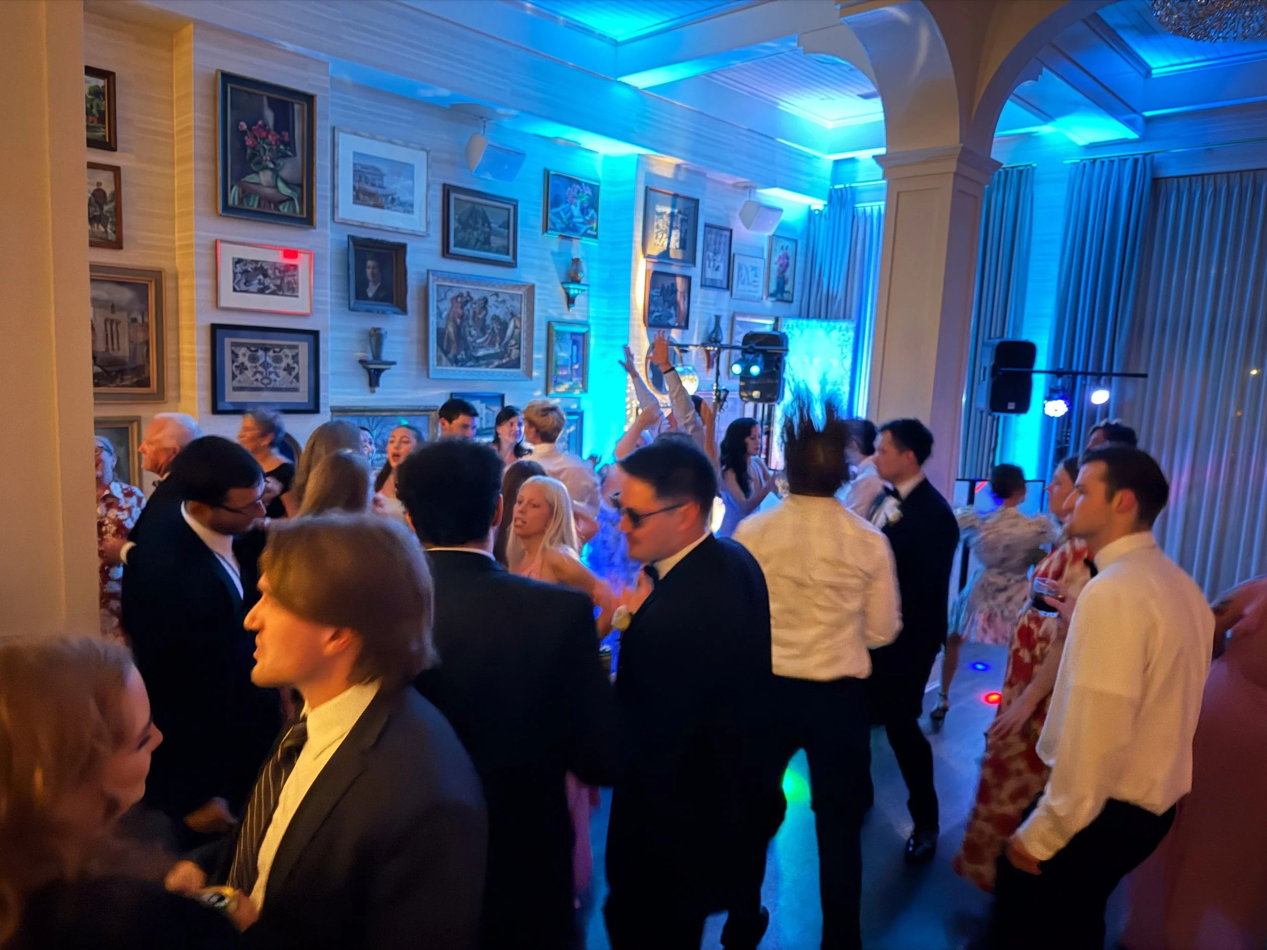 People dressed in formal attire dancing and socializing at a party in a well-decorated room with framed artwork on the wall and blue uplighting.