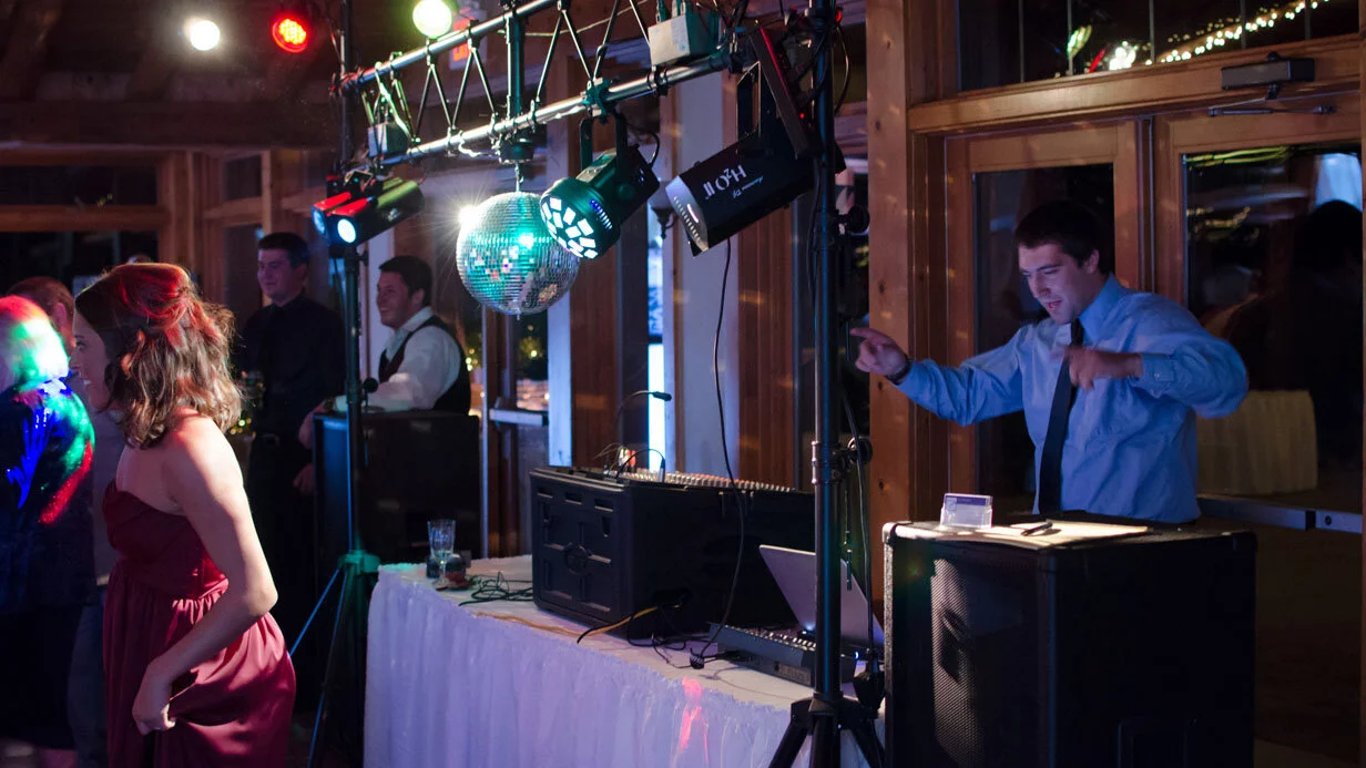 Indoor party scene with a DJ in a blue shirt playing music near a table with lighting equipment and a disco ball, with people dancing and socializing nearby in dim lighting.
