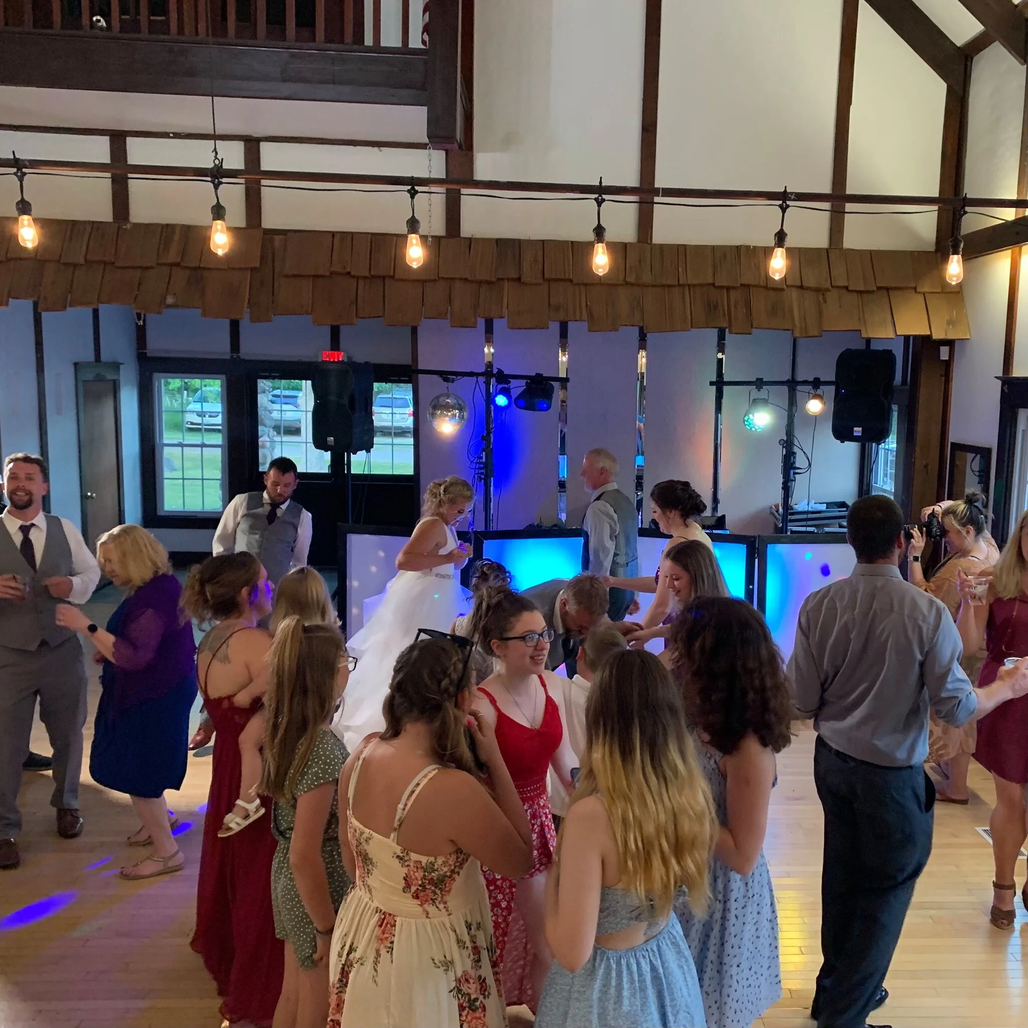 A group of people dancing at a wedding reception indoors, with a bride in a white dress visible. The venue has a wooden interior and string lights hanging from the ceiling. A DJ booth and sound equipment are set up at the back.