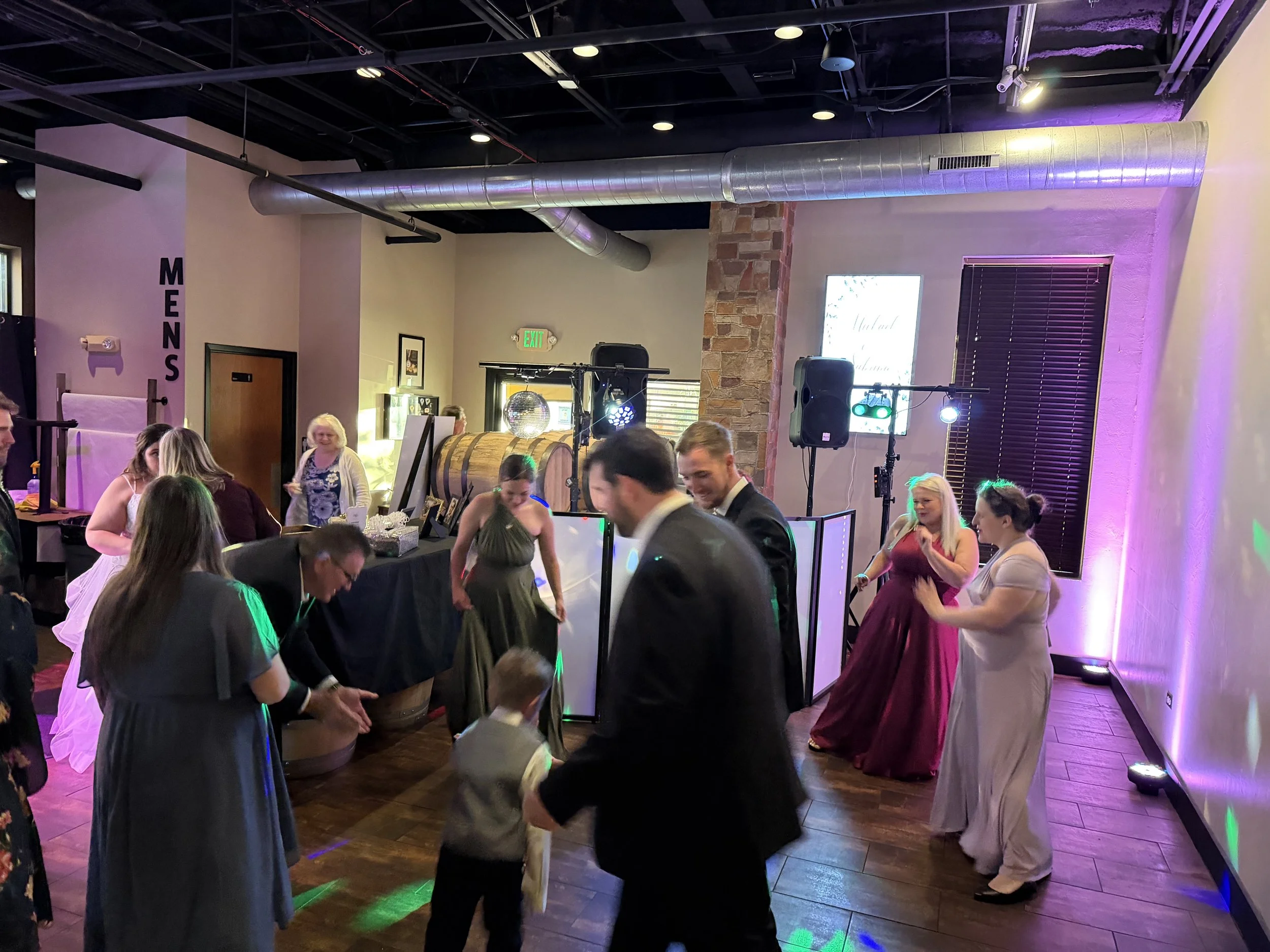 People dressed in formal attire dancing and socializing at a party with colorful lighting, wooden floors, and a DJ setup with speakers.