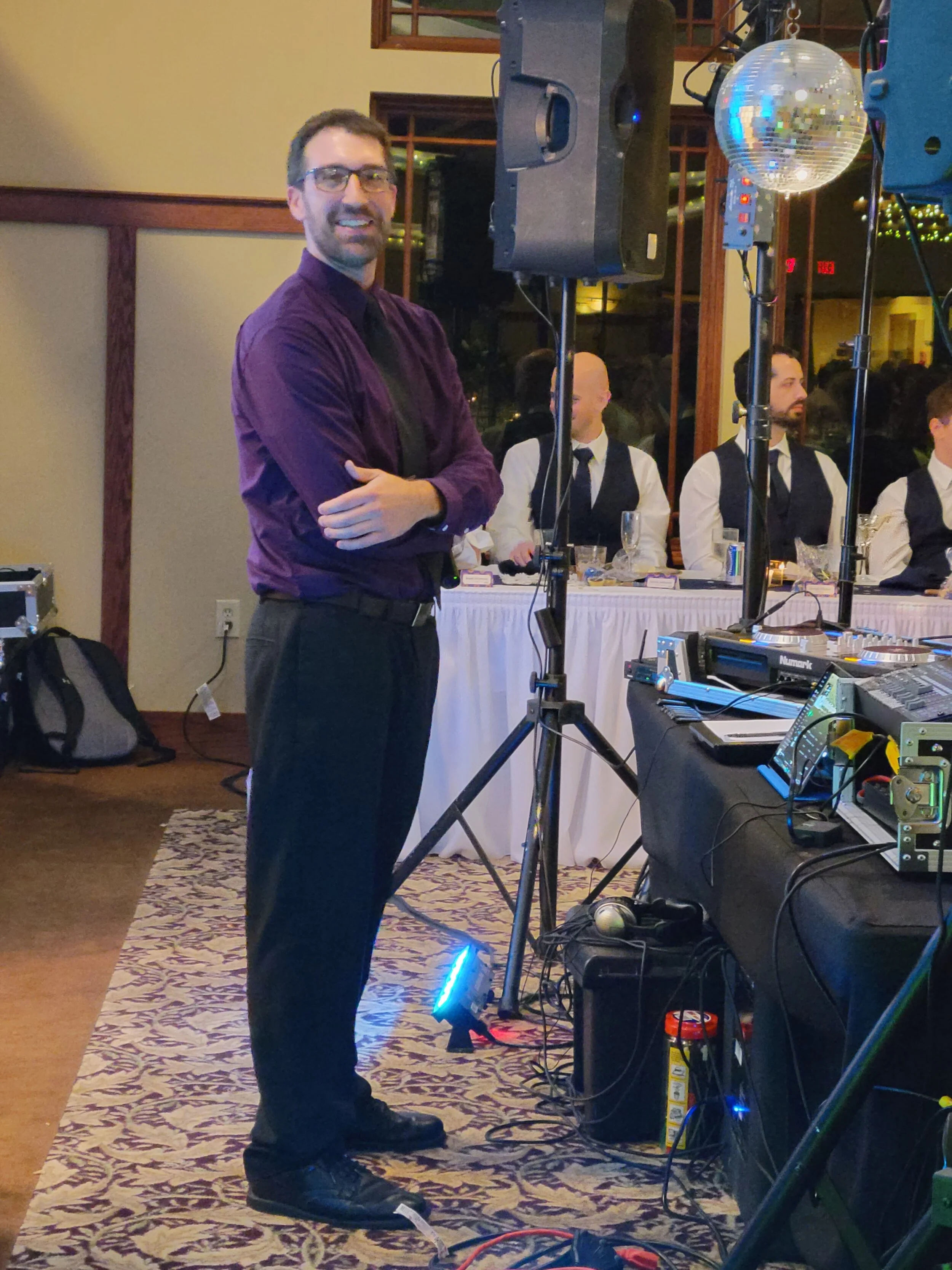 Man in purple shirt at event with DJ equipment and disco ball