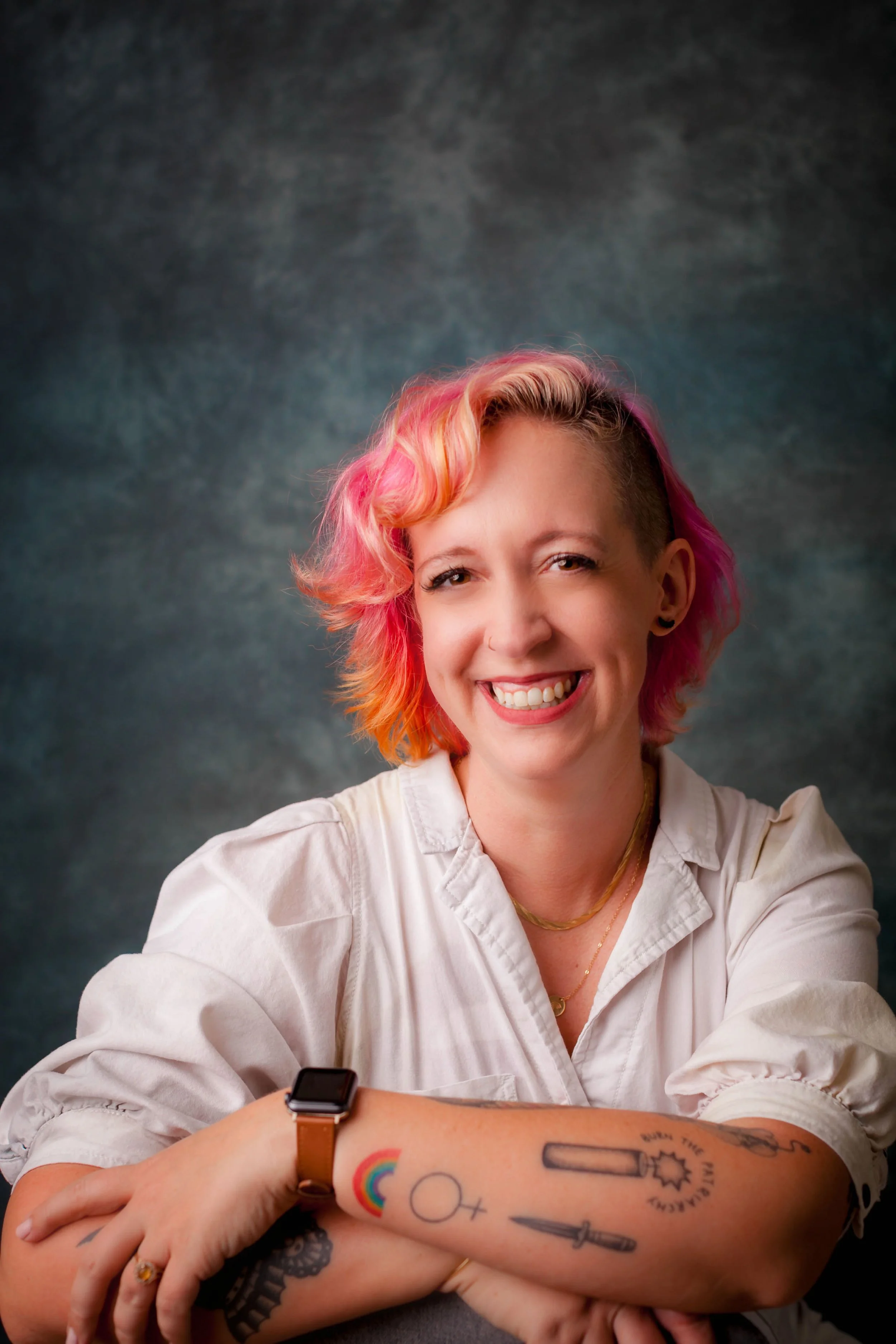 A smiling woman with pink and orange hair, wearing a white shirt, sitting with her arms crossed, showing colorful tattoos on her forearm including a rainbow, a female symbol, and a dagger, against a gray textured background.