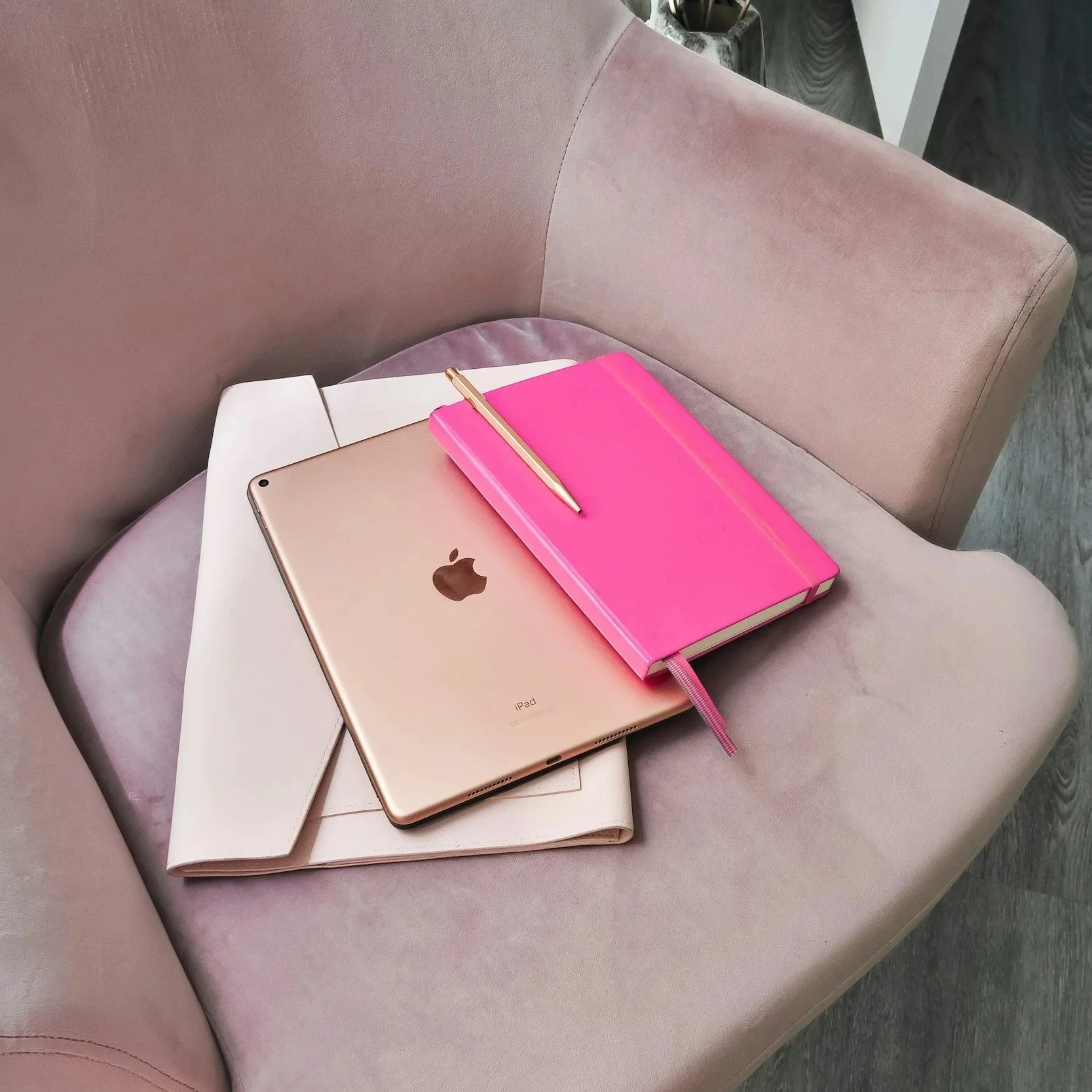 An iPad, a pink notebook with a gold pen, and another notebook partially covered by a white folder on a soft pink armchair.