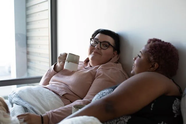 two plus-sized women lay in bed. one is caucasian with black rimmed glasses, a blush hoodie and short hair, holding a coffee cup. The woman lying by her is Black with natural hair, and is also holding a coffee cup. They are grinning at each other.