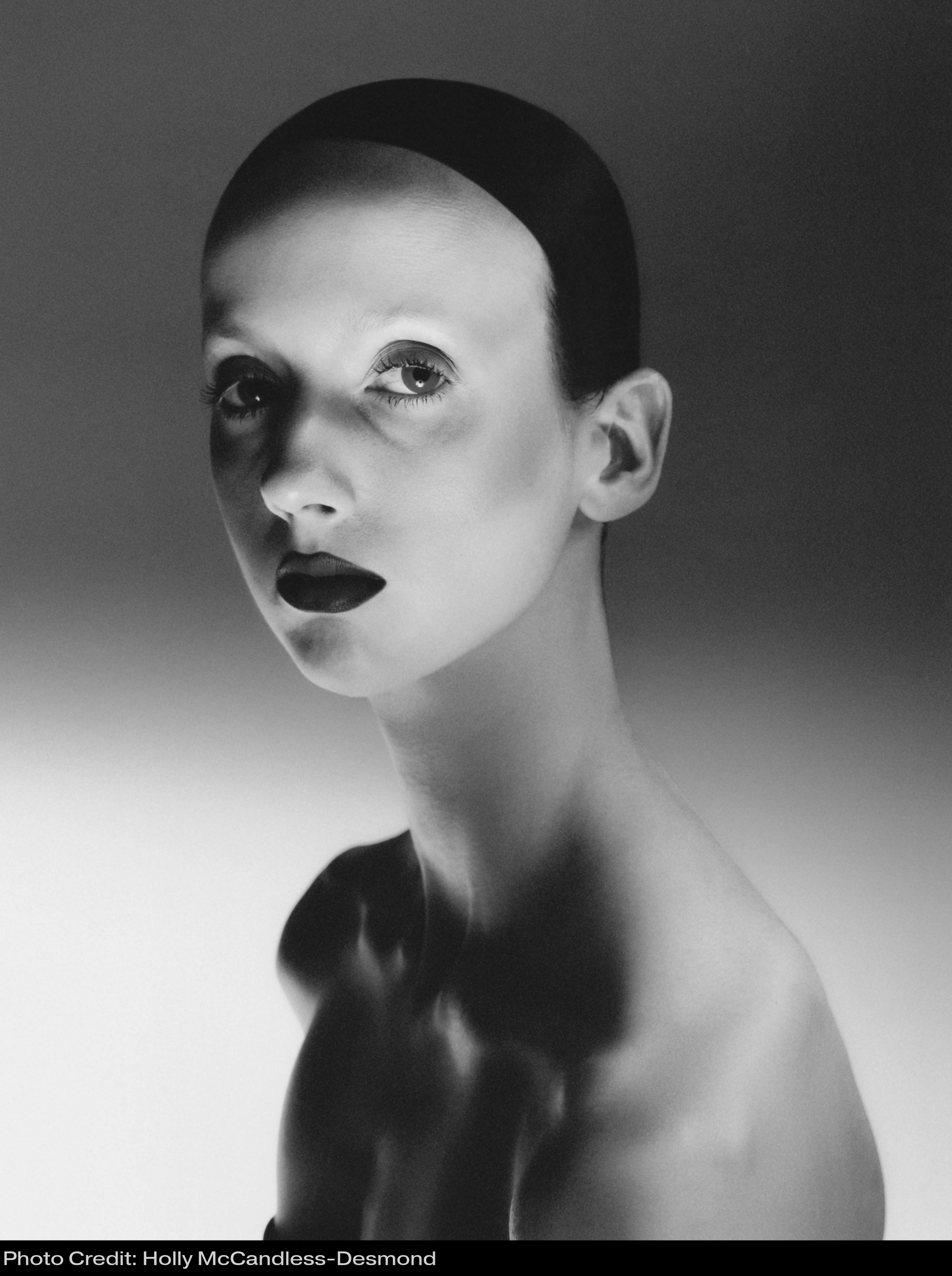 A head shot of Zara in black and white. She has pale skin and light eyebrows with dark slicked back hair. She looks at the camera.
