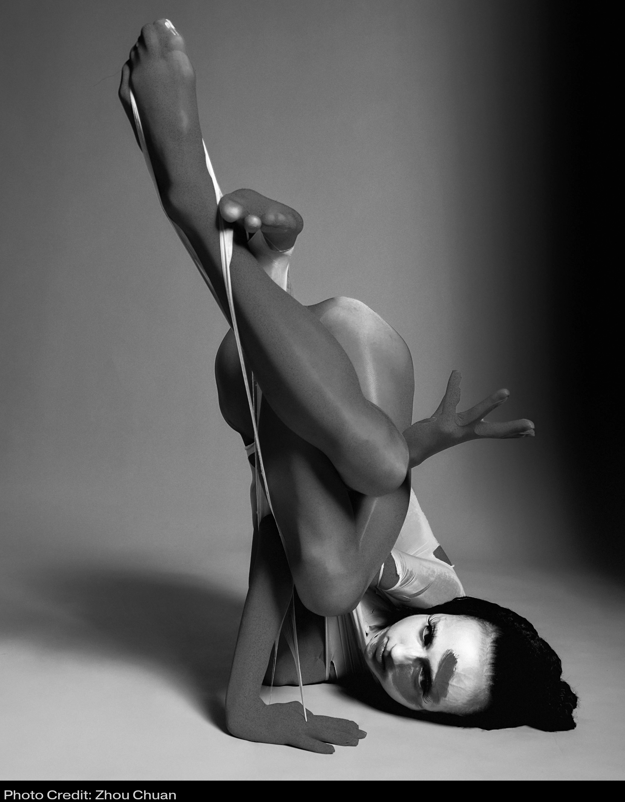 Zara balancing on her shoulder with her body curled into a sculptural shape with limbs intertwined. The image is black and white and she wears a catsuit and face paint with a painted squiggle shape across her face.