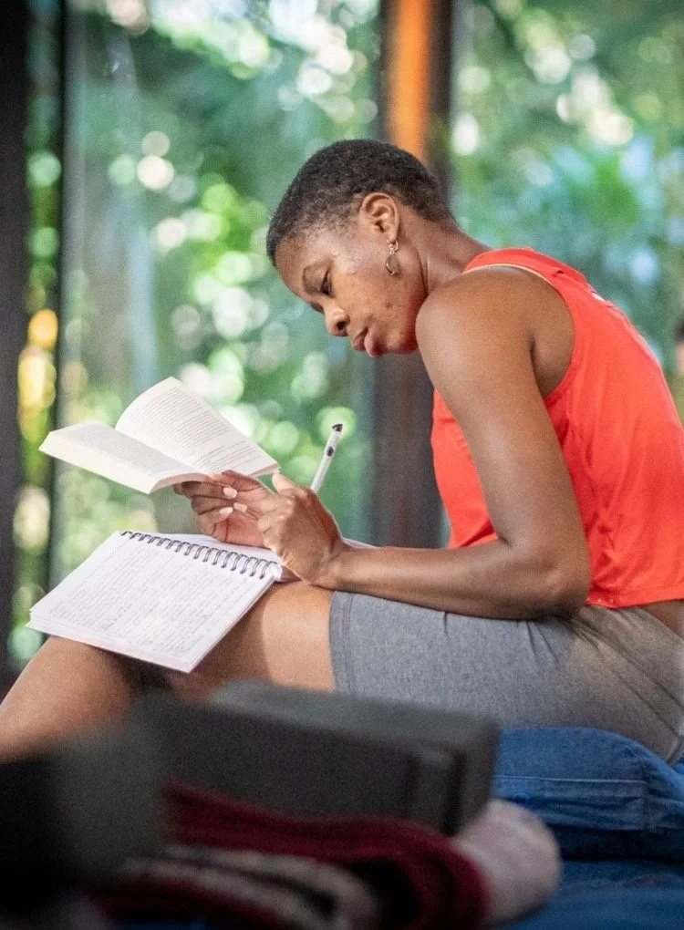 Medium brown skin woman wearing a red shirt and gray shorts is holding a pencil in one hand and a book in the other hand. A journal is open and resting on her knee.
