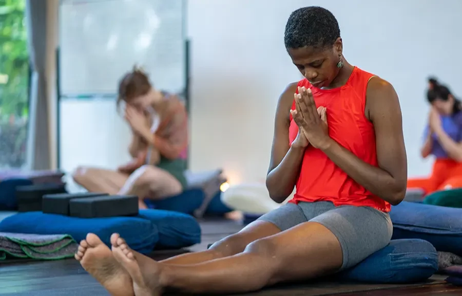 A brown skin woman wearing a red tank top and gray shorts is sitting on a  blue cushion bowing with her hands in prayer position at your heart