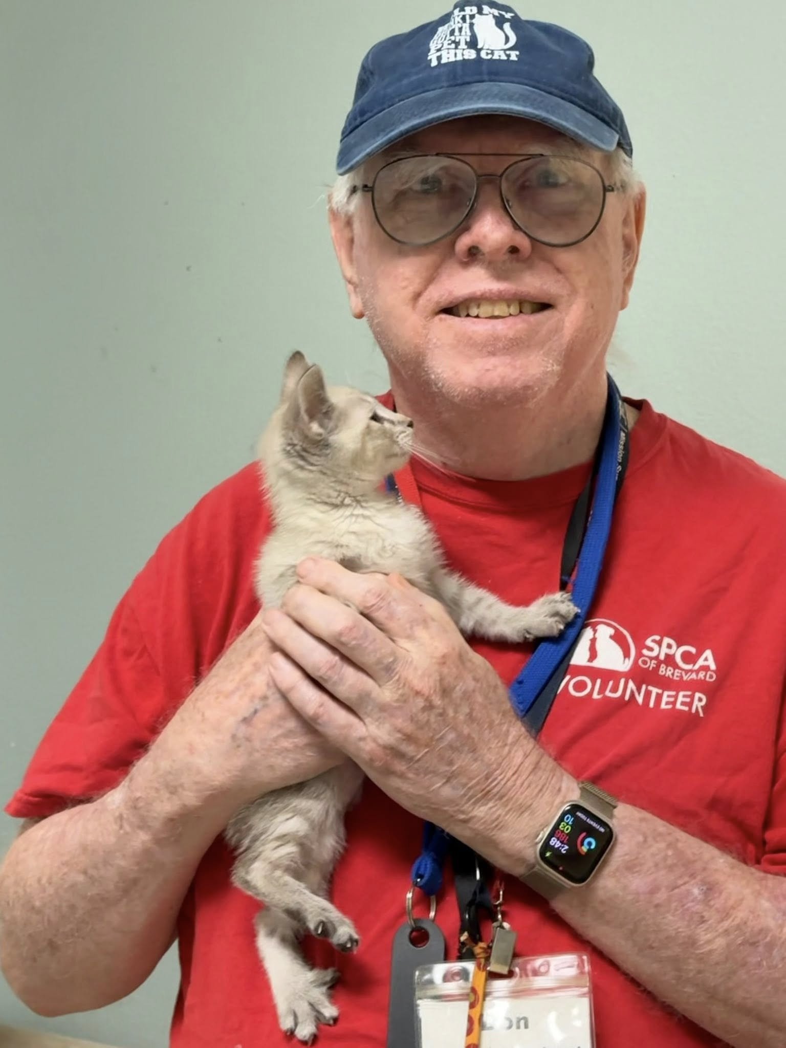 An older man wearing glasses, a blue cap with a cat graphic, and a red SPCA volunteer shirt, smiling while holding a kitten.
