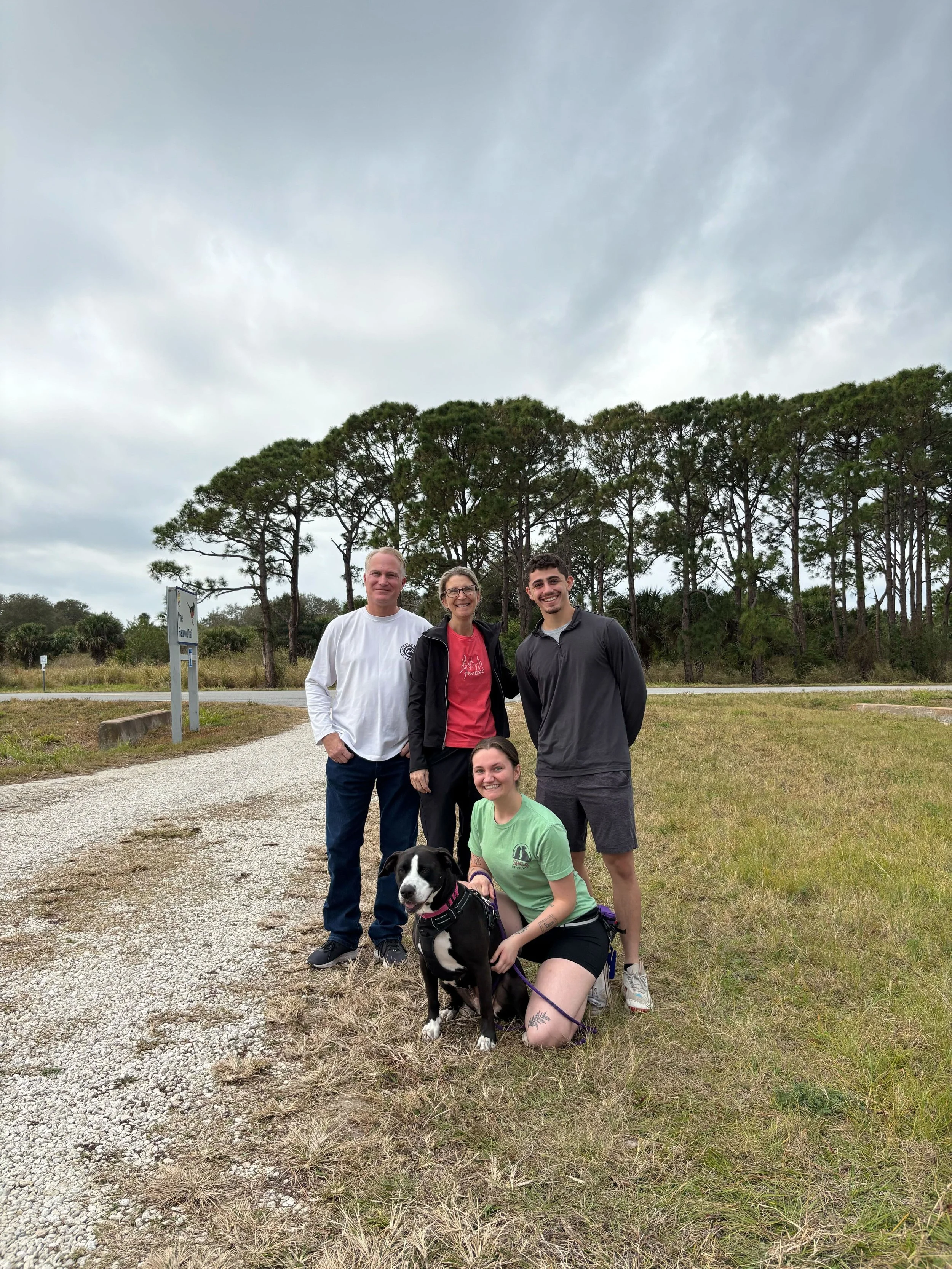 A group of four people and a black and white dog outside on a cloudy day, standing near a gravel path with trees in the background.