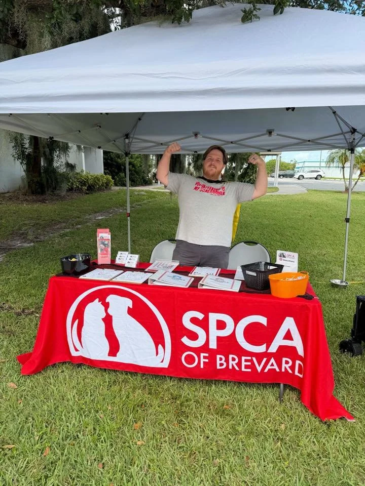 Man at an outdoor event booth for SPCA of Brevard, standing under a white canopy, flexing his arms, with a red tablecloth displaying the SPCA logo and name, surrounded by pet-related informational materials.