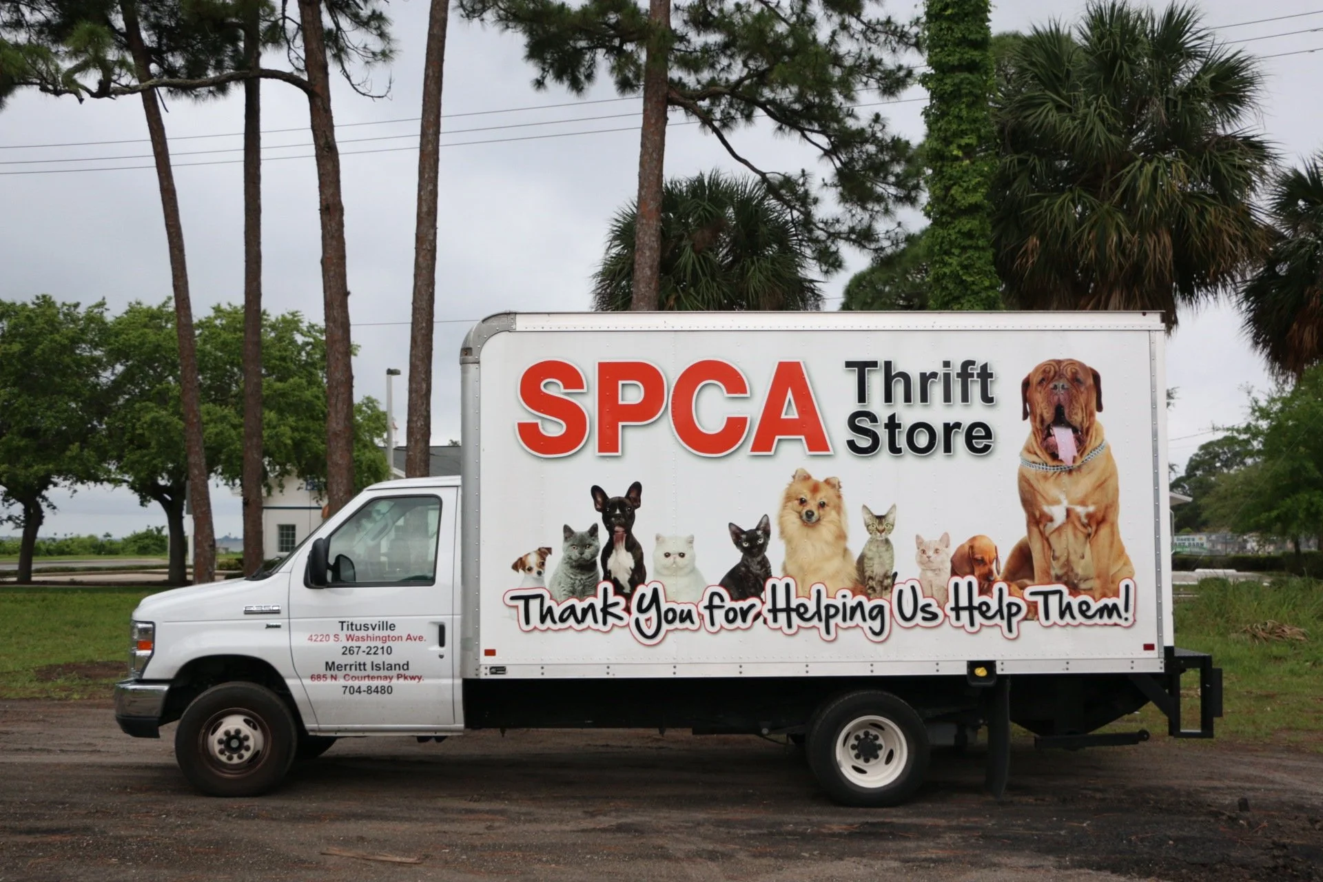 A white truck with a large sign on the side advertising an SPCA thrift store, featuring images of cats and dogs, and a sentence thanking customers for helping animals.