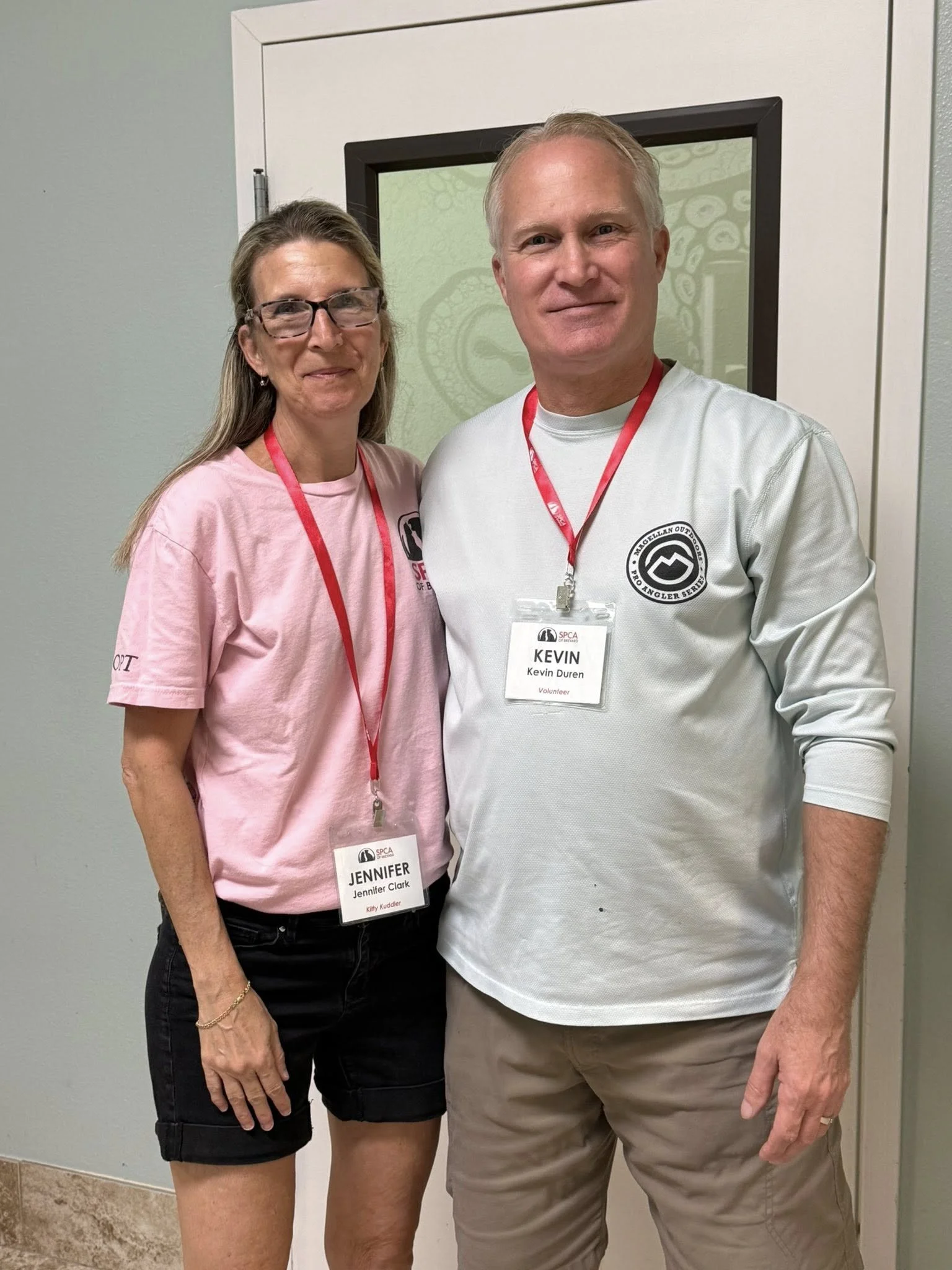 A woman and a man standing together, both wearing volunteer badges, smiling in front of a door with a decorative glass window.
