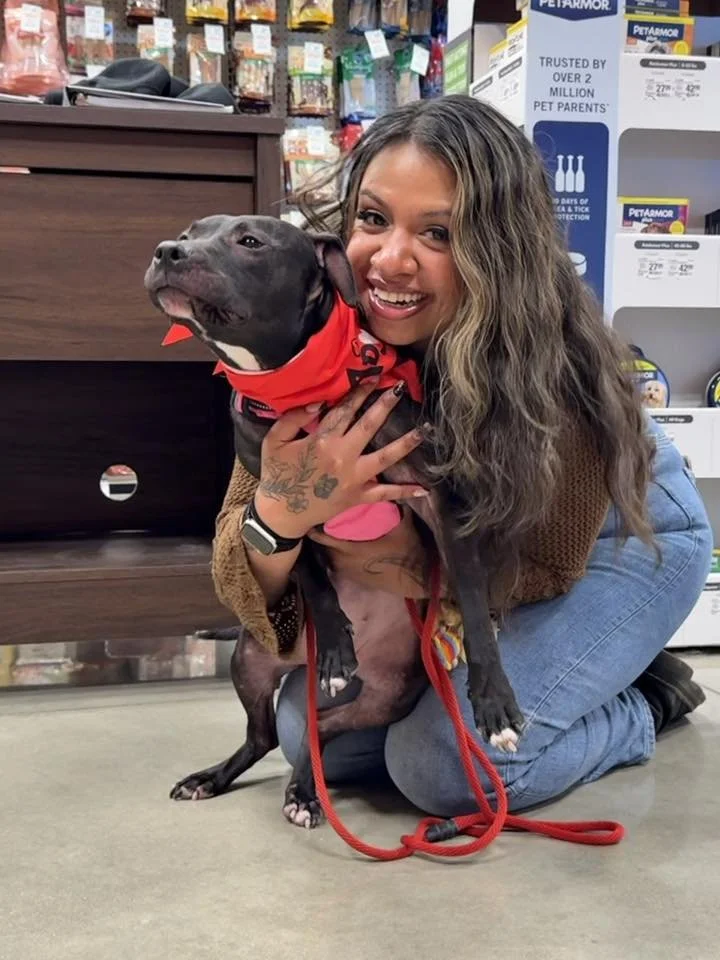 A woman with long wavy hair smiling and kneeling on the floor, holding a black dog with a red bandana around its neck in what appears to be a pet store.