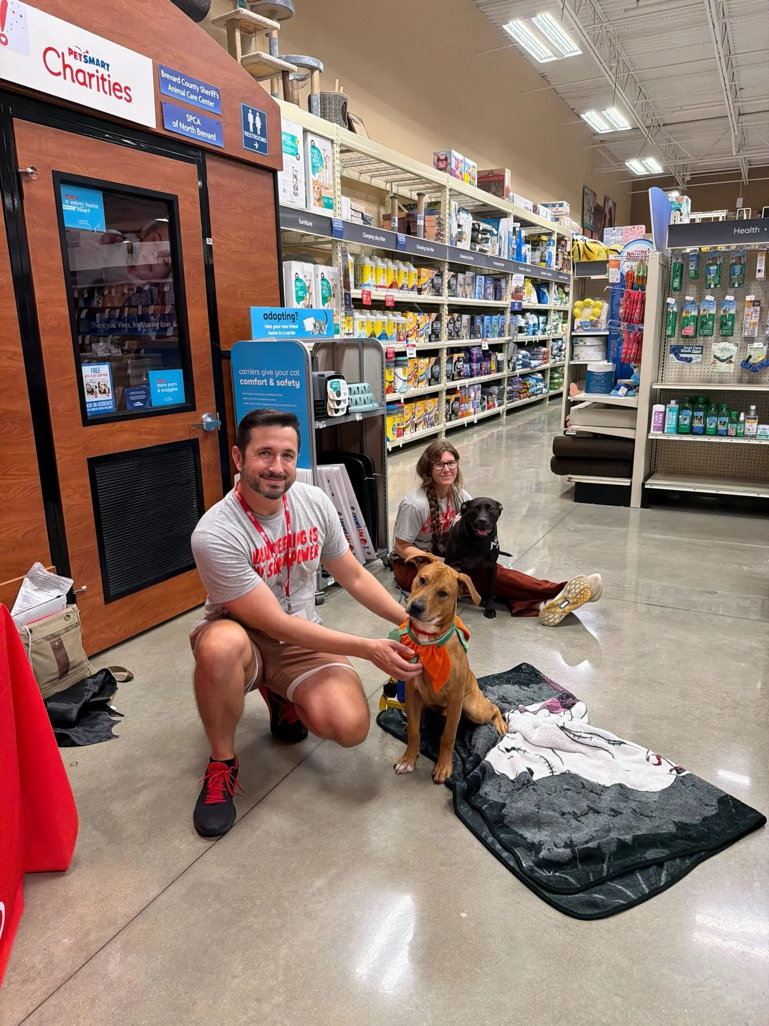 Two people with two dogs sitting on the floor inside a pet store near the entrance, with shelves of pet supplies in the background.