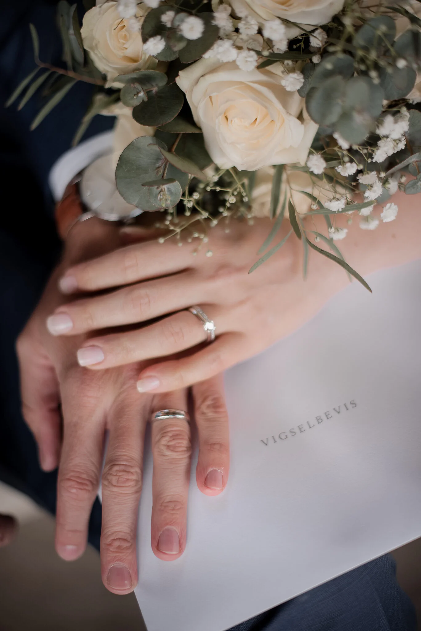 Ett bröllopspar visar sina ringar, med en blomsterbukett, på en vit bakgrund med texten 'VIGSELBEVIS'.
