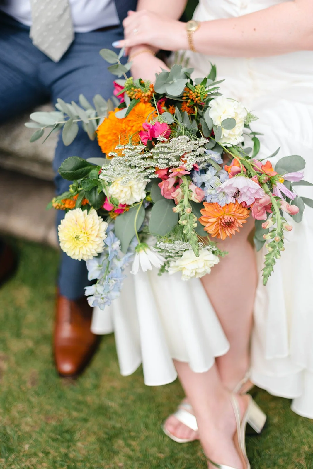 Brud som håller en stor blomsterbukett med färgglada blommor, inklusive orange, rosa, vit och gröna blad, medan en person i kostym står bredvid.