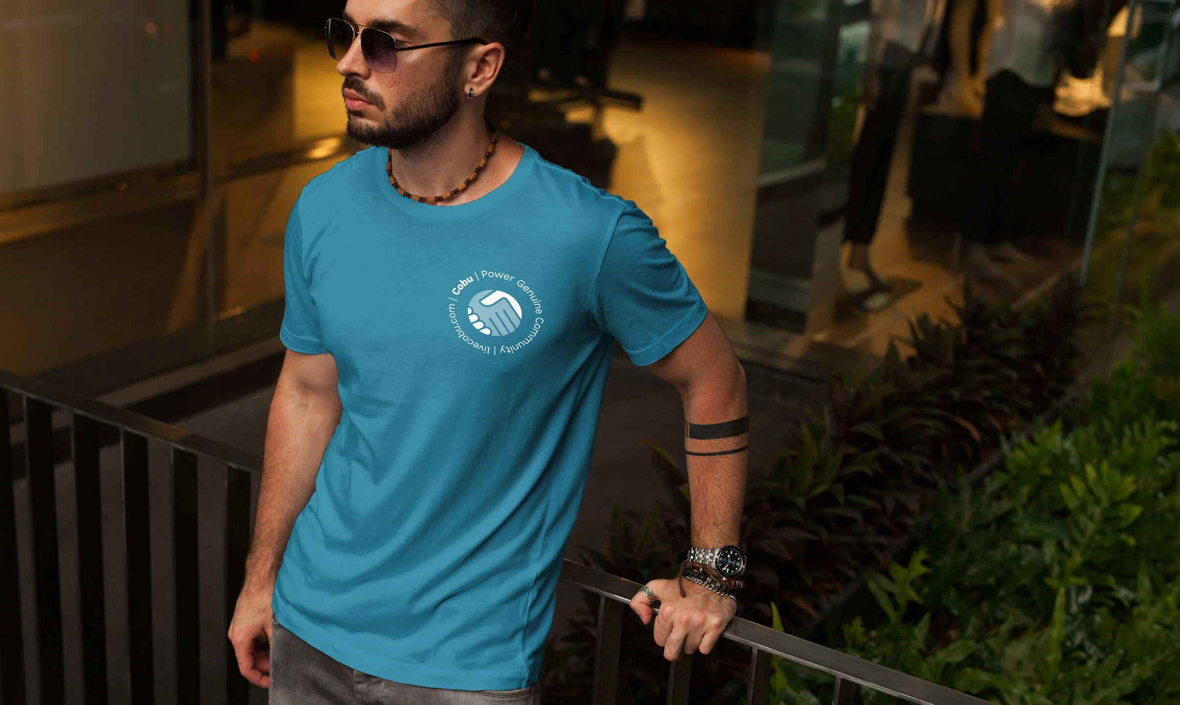 A young man with tattoos and sunglasses, wearing a blue Cobu t-shirt, standing outdoors at night, holding a railing, wearing jewelry including a beaded necklace, watches, and rings.