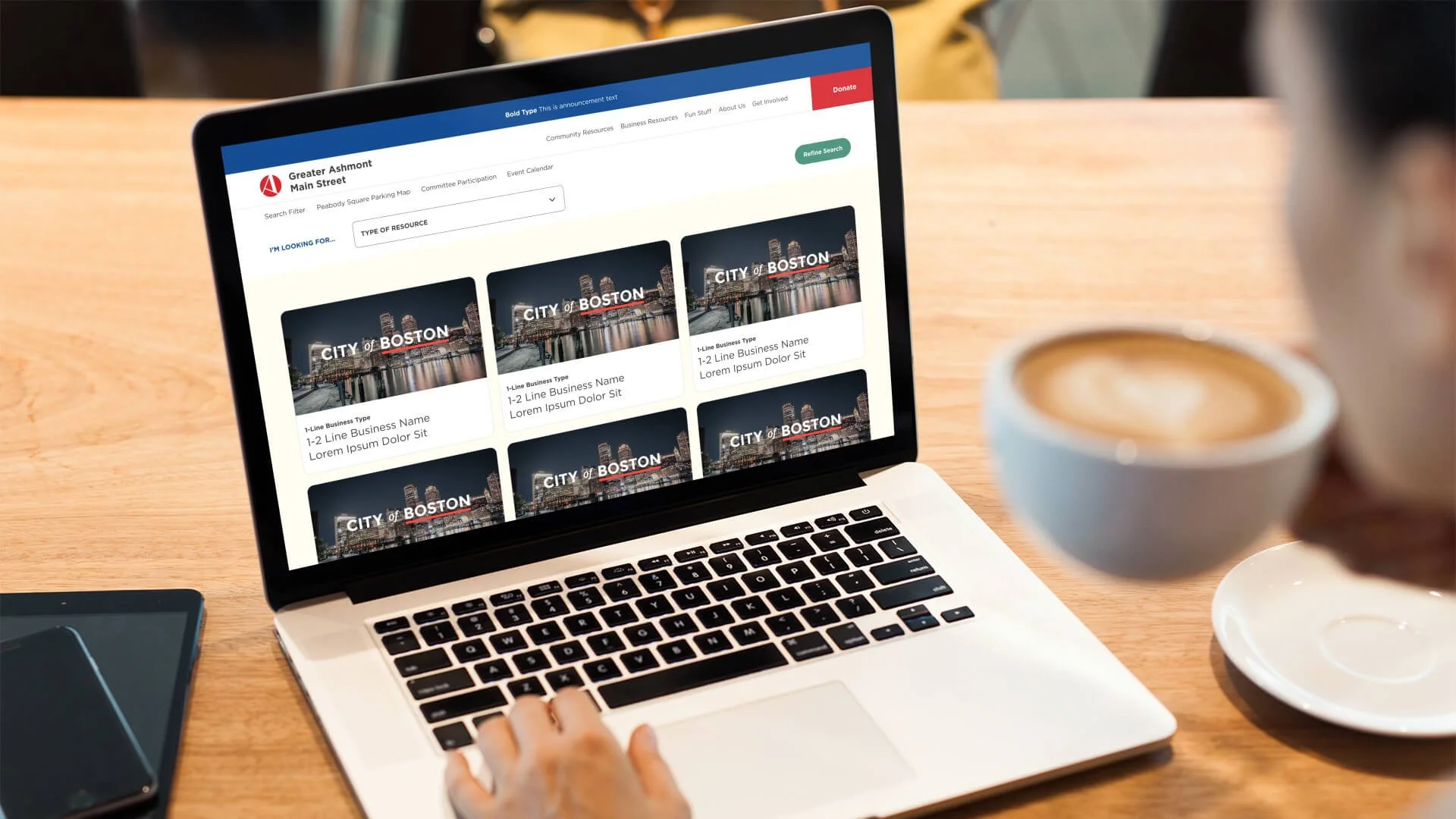 A laptop on a wooden table displaying a city planning website for Boston with resource listings and a green refine search button, a coffee cup to the right, and a smartphone to the left, with a person in the background taking the photo.
