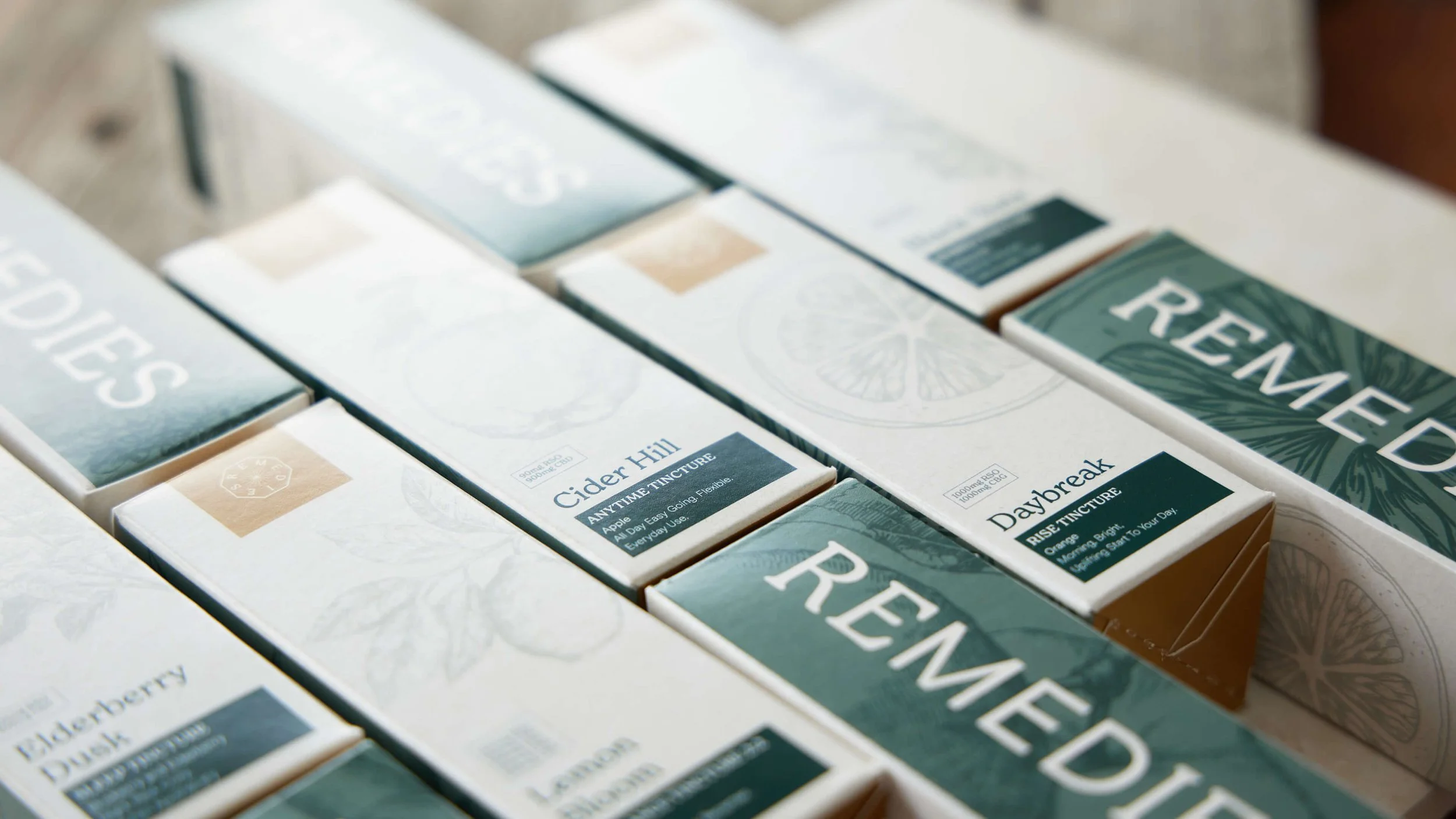 Multiple boxes of Remedies tinctures displayed on a surface, featuring flavors like Cider Hill and Daybreak, with natural illustrations and labels.
