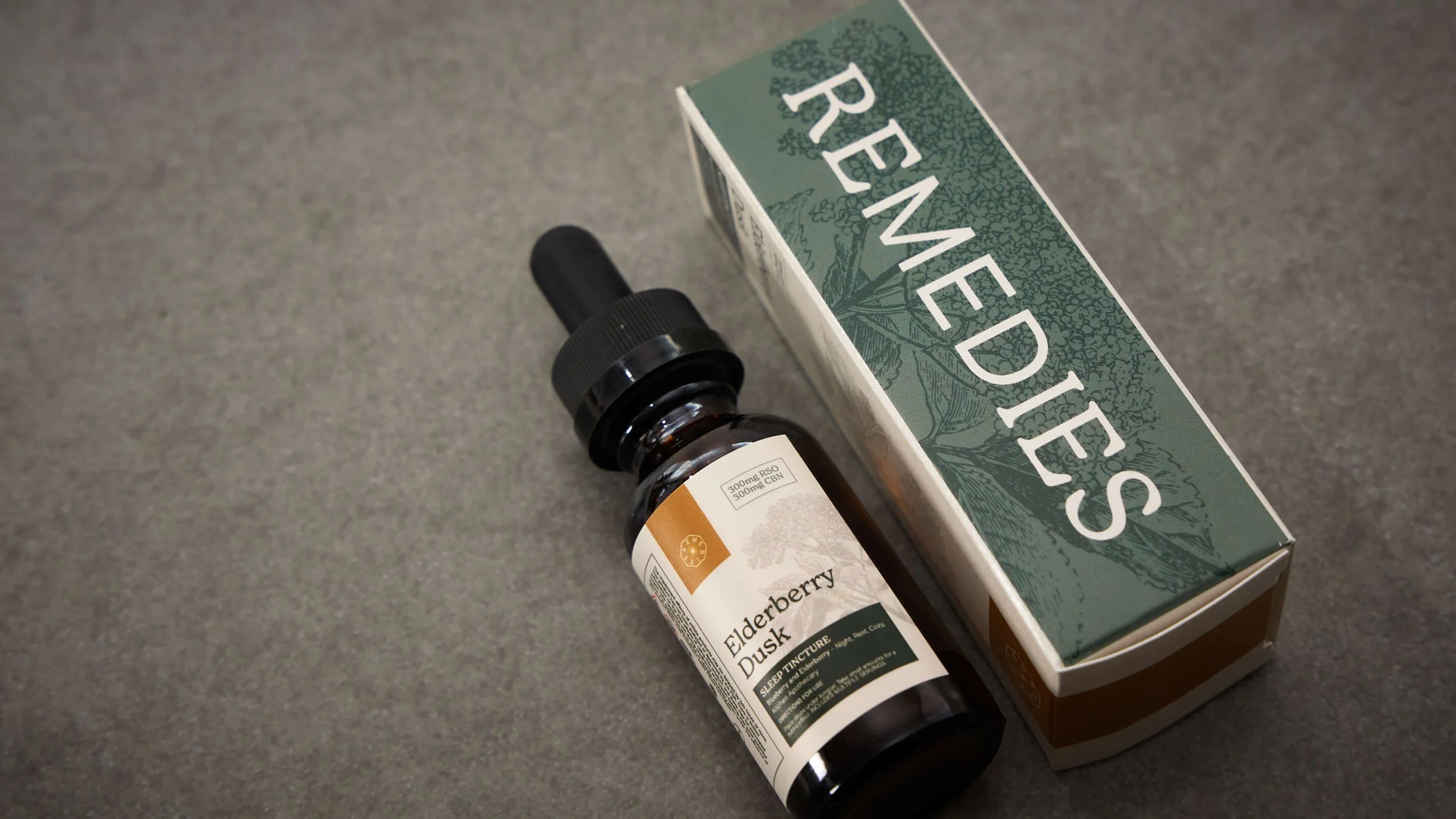 A box labeled 'Remedies' with a drawing of a botanical on it, next to a small dark brown tincture bottle with a white label and black cap, containing elderberry syrup.