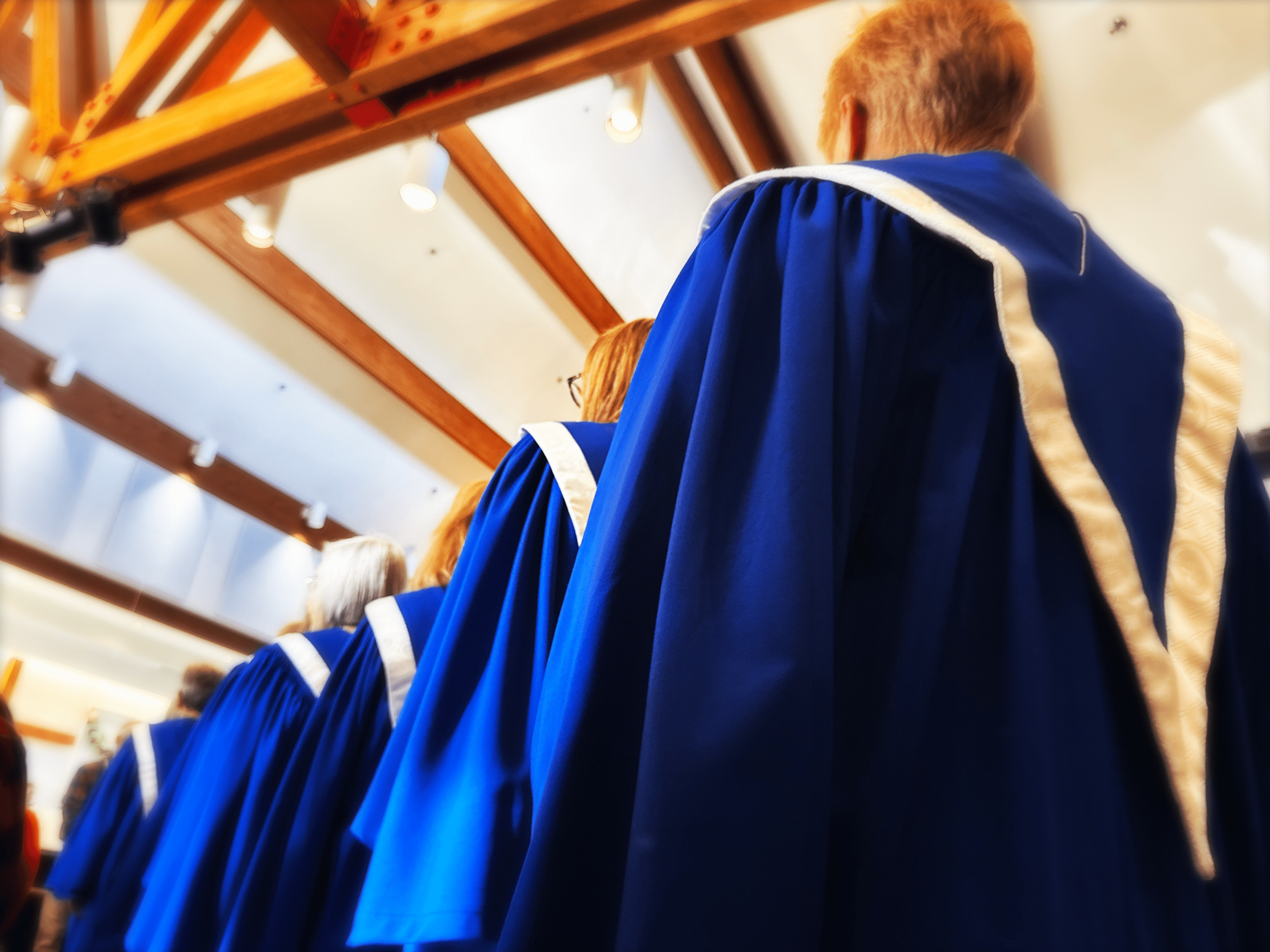 A group of elderly women wearing blue choir robes with white stoles and crosses on the chest, singing and holding red hymn books during a church service or choir performance.