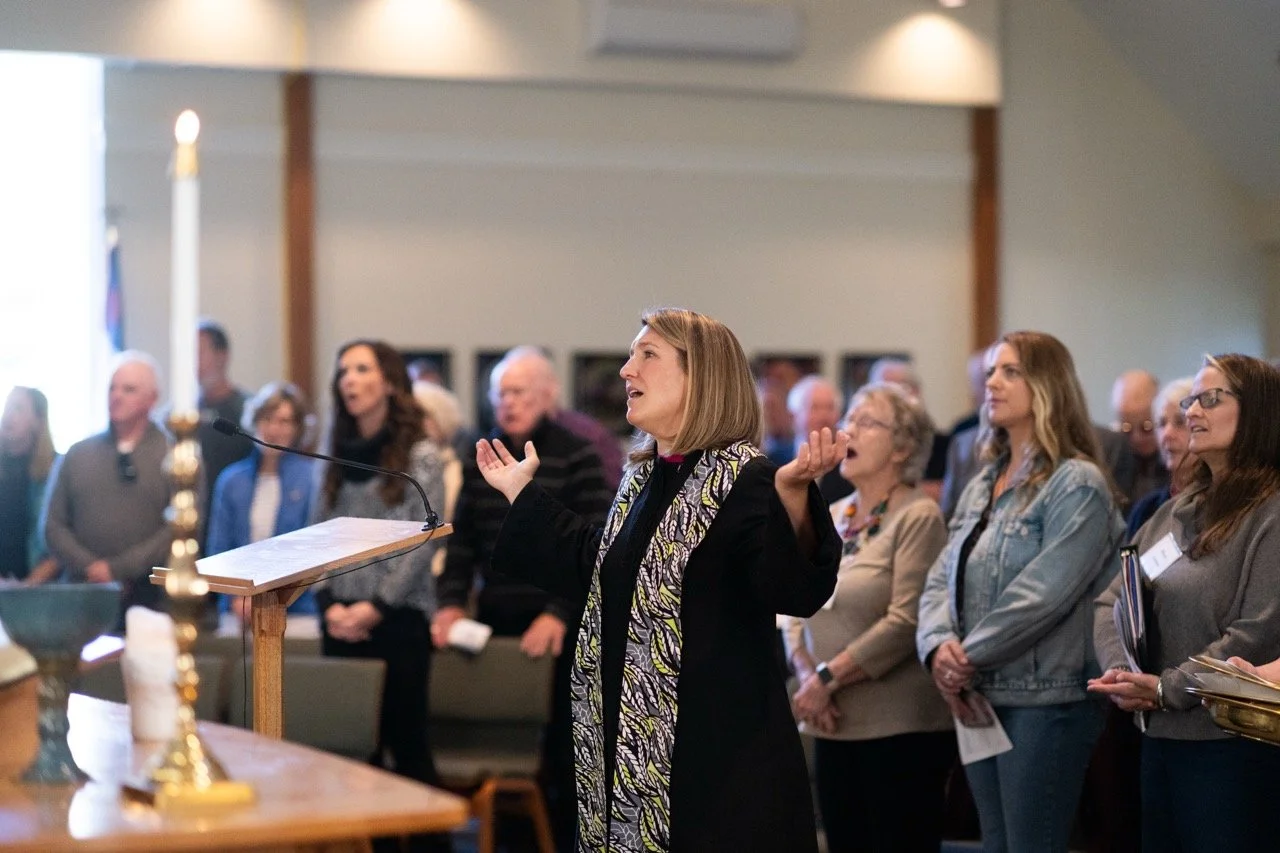 People attending a church service, including a man in a graduation gown and a woman holding a book, standing with eyes closed in prayer.