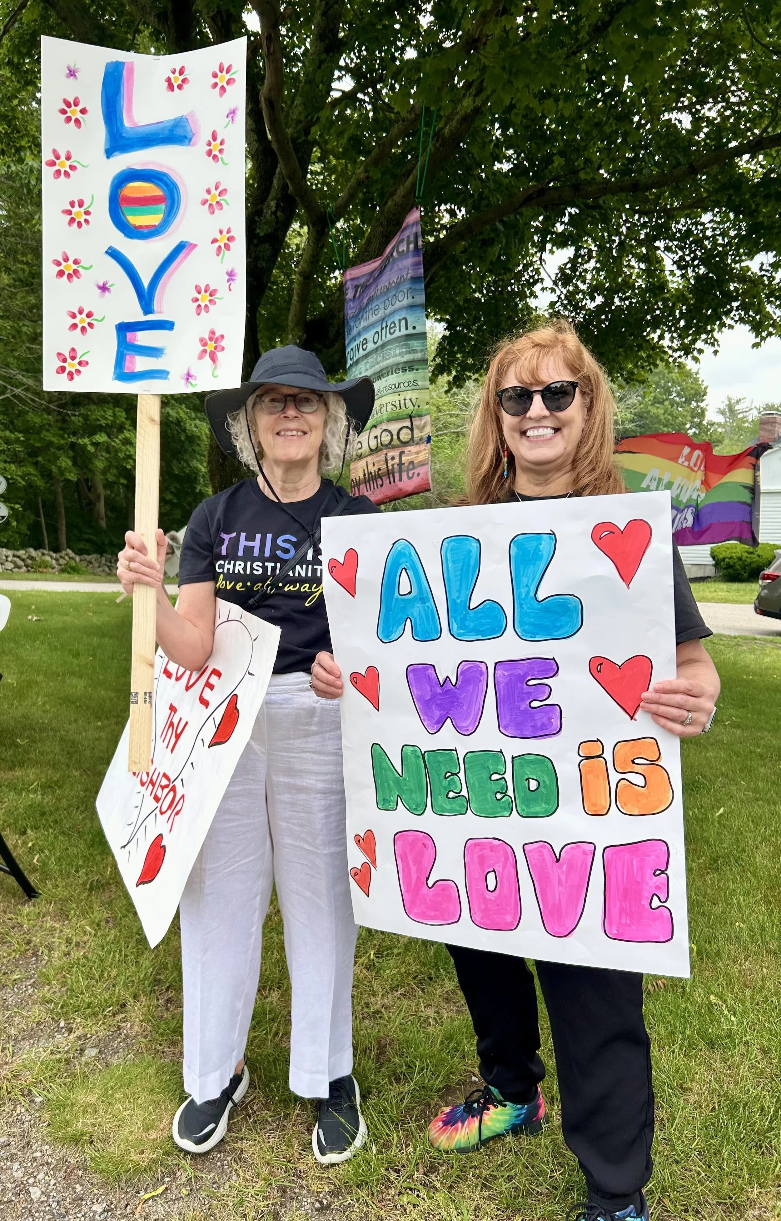 Two women smiling outdoors at a pride event, holding handmade signs with messages of love and inclusion. One sign says "LOVE" with colorful flowers, and the other says "ALL WE NEED IS LOVE" with colorful hearts.