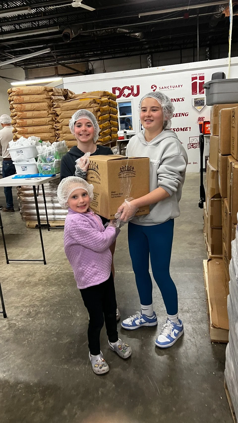 three people wearing hair nets and hats standing in front of boxes of rice and pasta pose for a picture, smiling
