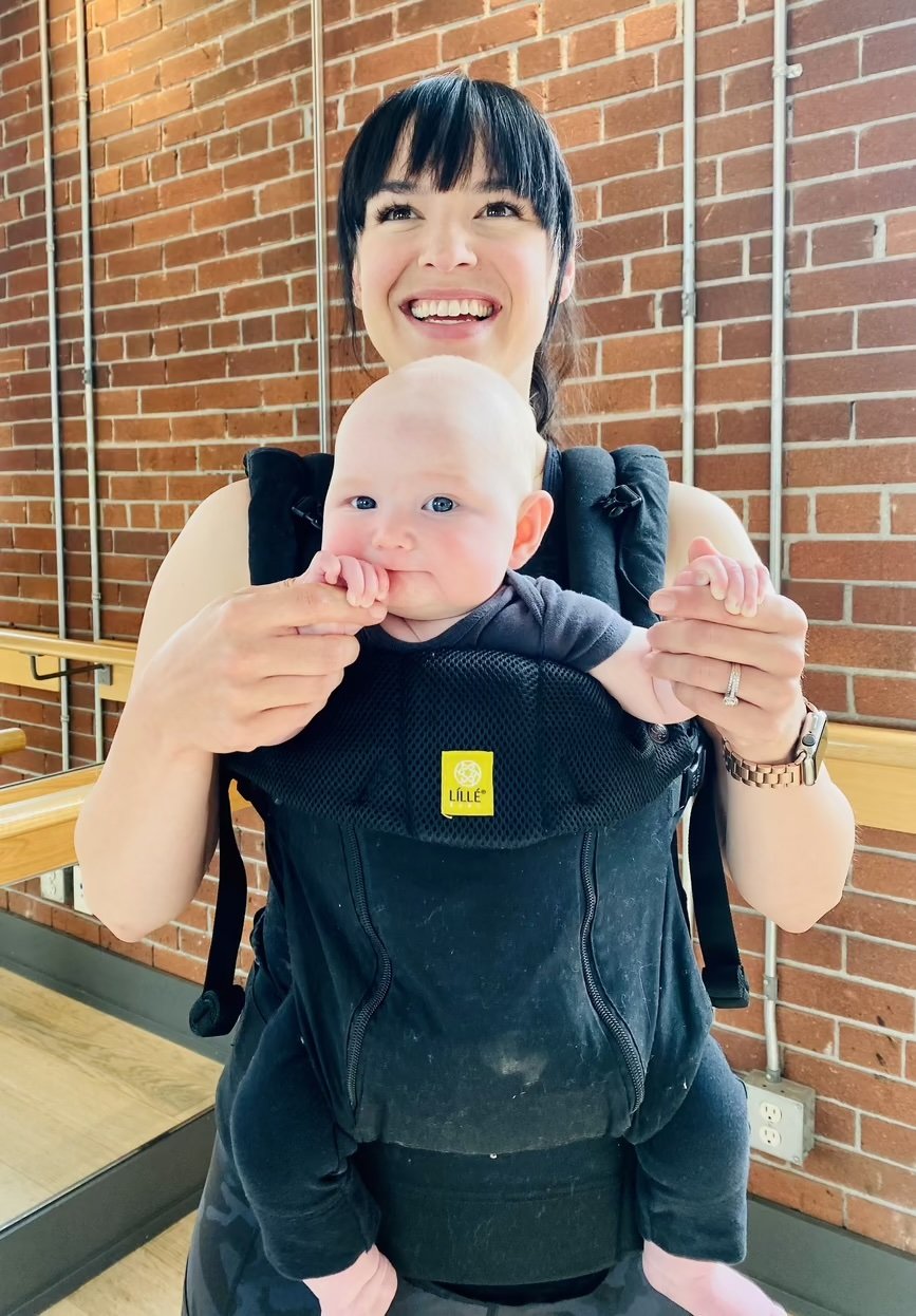 A smiling woman wearing a baby in a front facing carrier in a fitness studio with brick walls and a ballet barre
