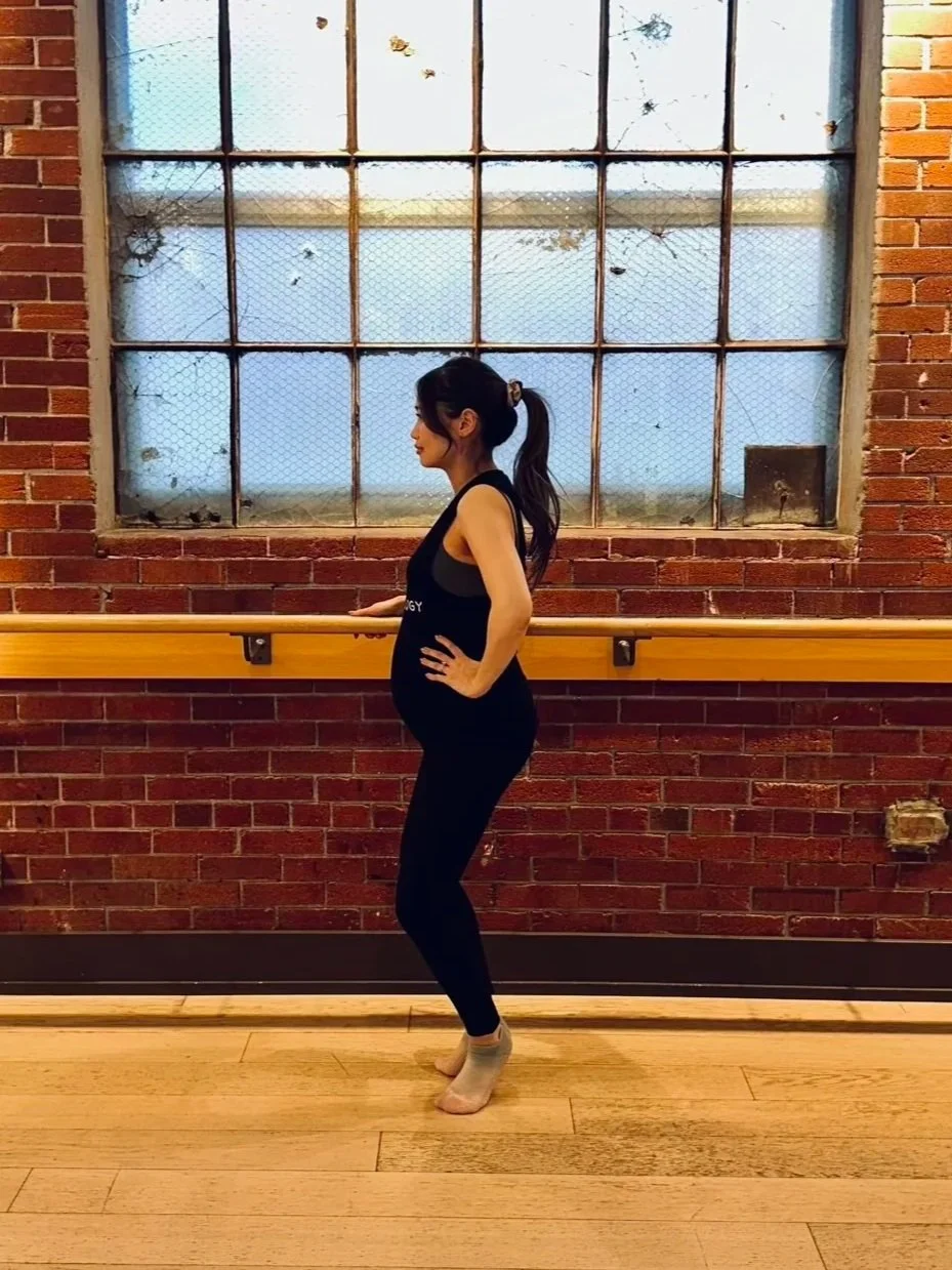 A pregnant participant at a ballet barre in fitness studio brick and a big window