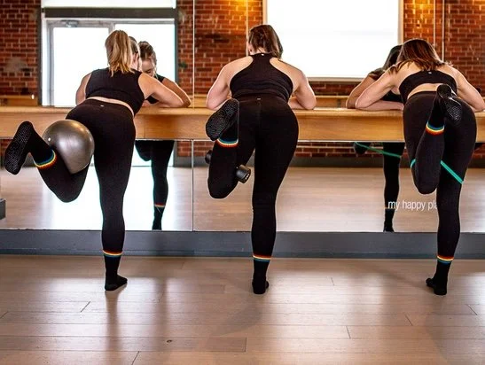 3 women wearing black activewear performing a barre exercise with different props in a bright fitness studio with large mirrors and windows, brick walls and a ballet barre.