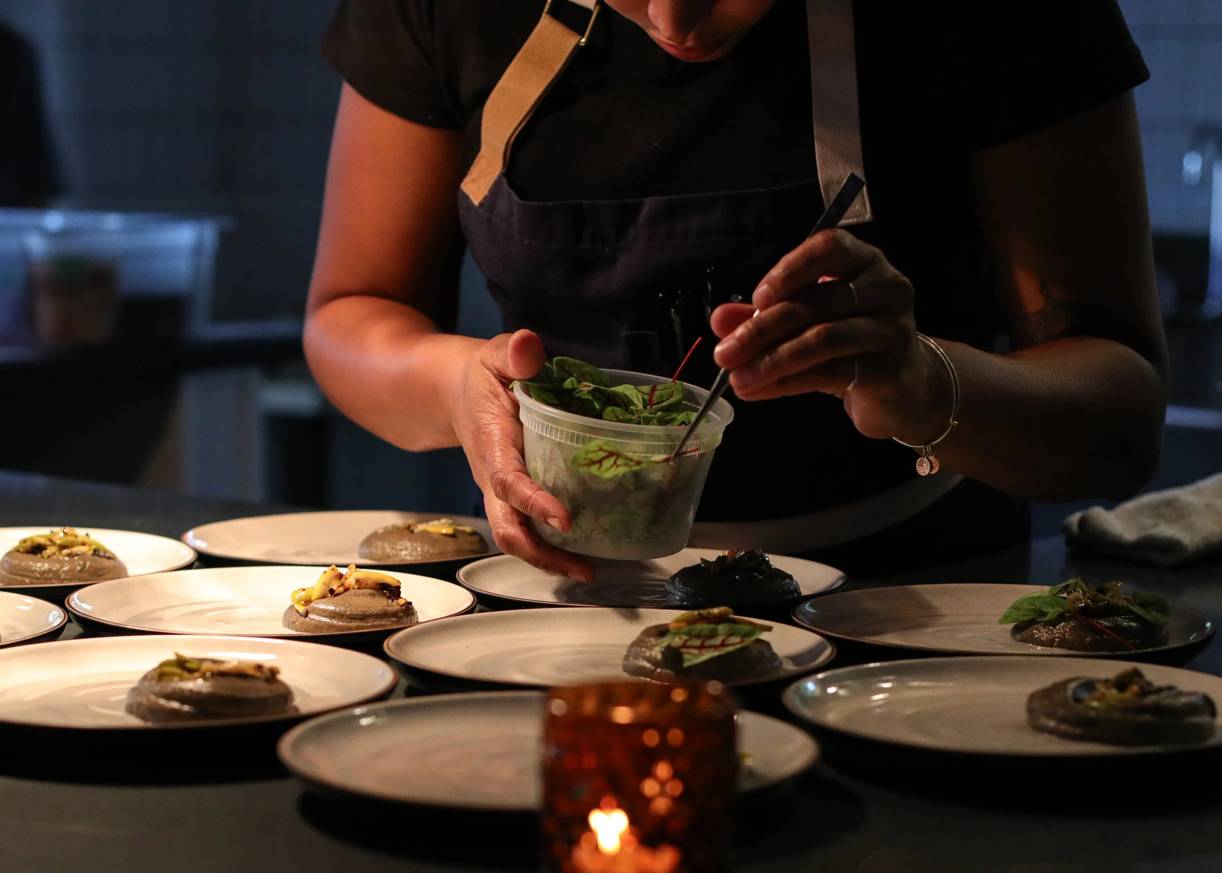 A person in a black shirt and apron is placing fresh greens into a clear plastic container, surrounded by plates of plated appetizers or desserts on a dark countertop, with a warm, dimly lit background.