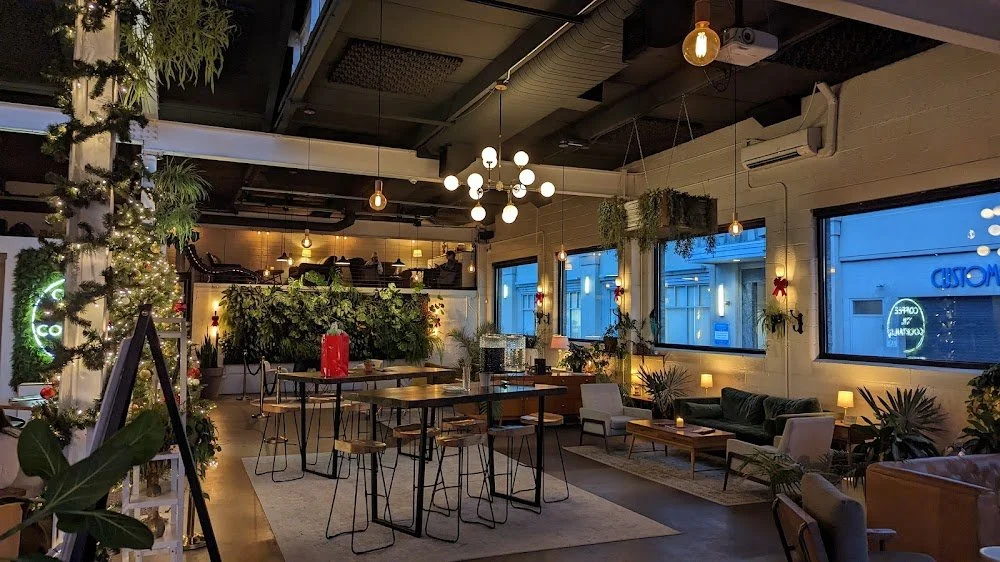 Interior of a cozy cafe or lounge decorated with Christmas greenery, string lights, and poinsettias, with seating areas, large windows, and warm lighting.