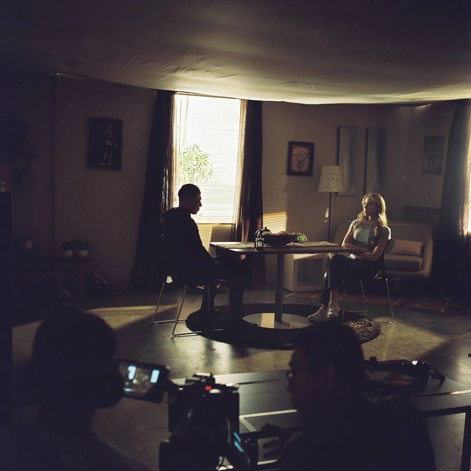 Two people sit at a table in a dimly lit room with natural light coming through windows. The scene appears to be a photo or video shoot, with a camera and crew members visible.