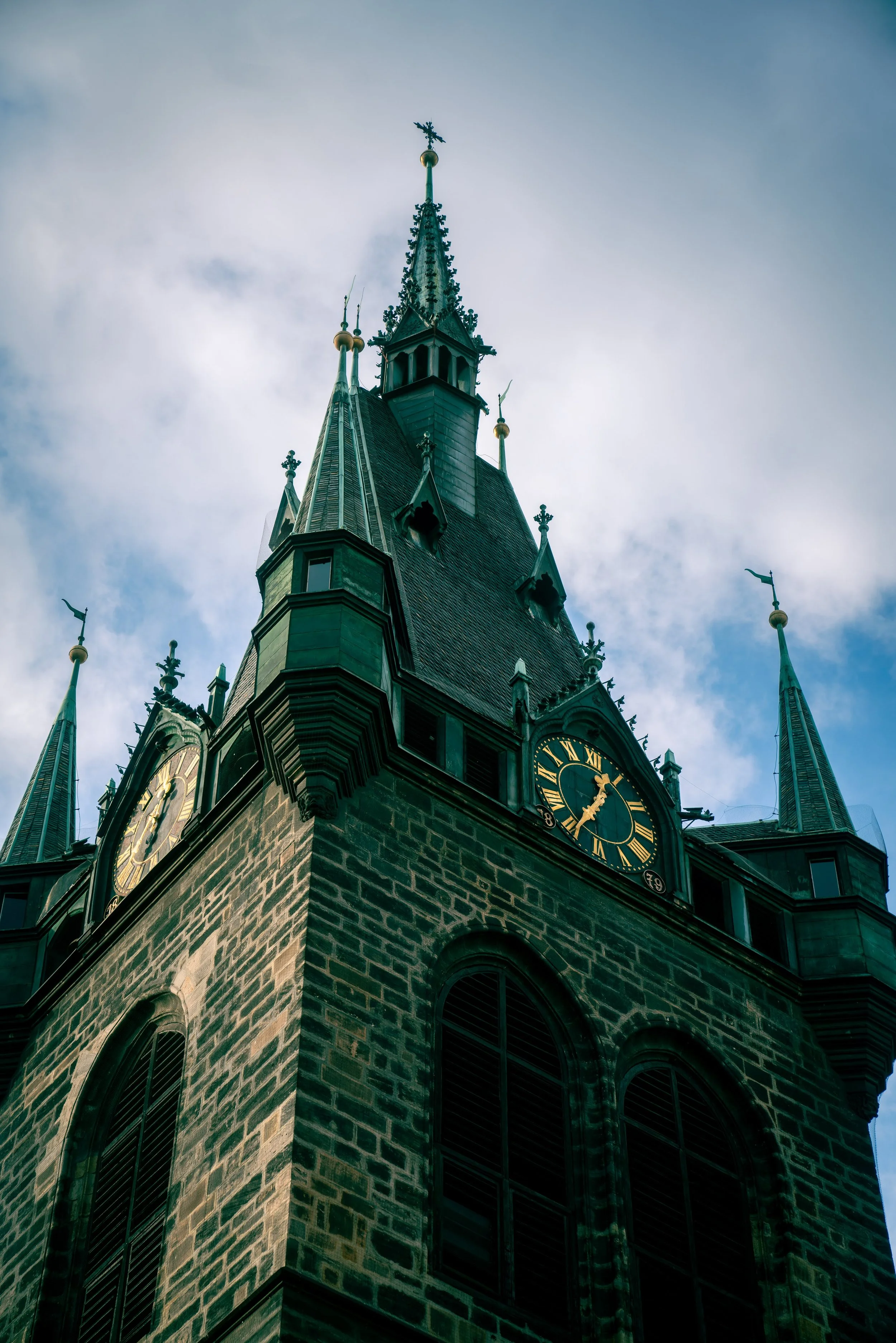 Close-up view of the tall clock tower of a historic Gothic-style church or cathedral with spires and a pointed roof against a cloudy sky.