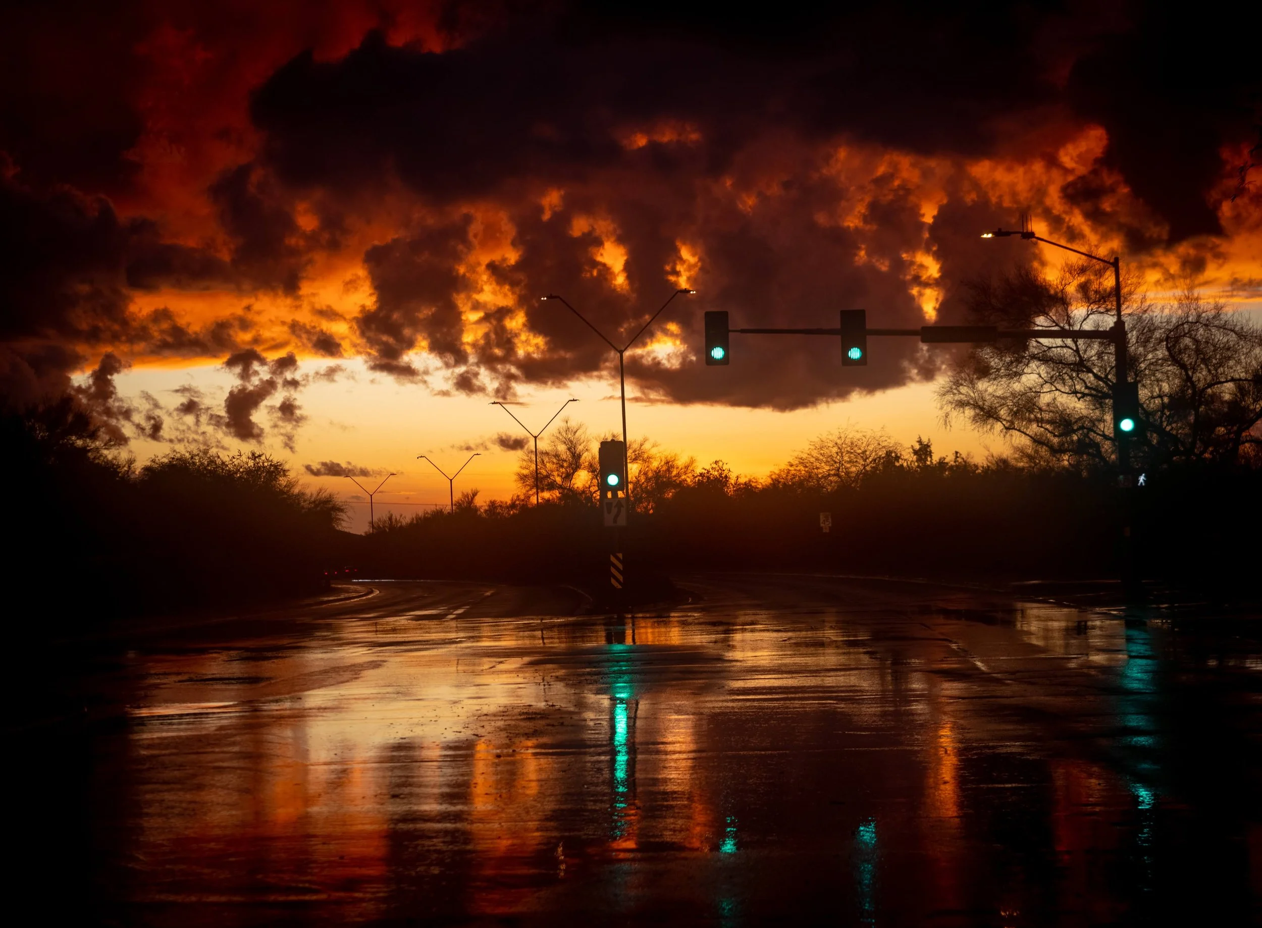 Sunset sky with dark clouds and illuminated traffic lights over a wet, reflective street with trees in the background.