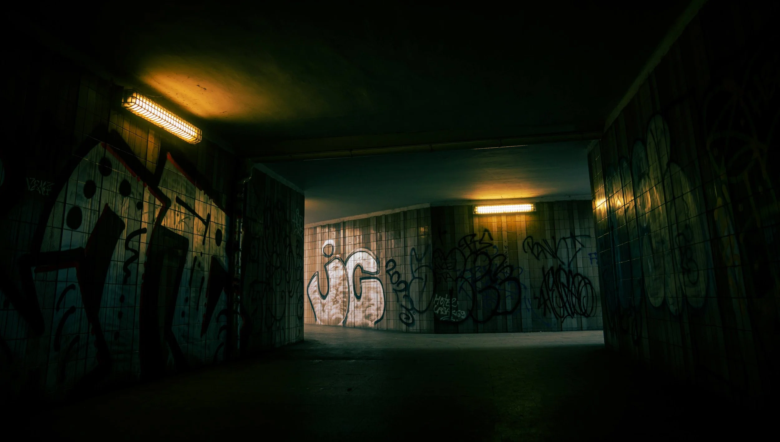Underground tunnel with graffiti on tiled walls, illuminated by overhead lights, featuring various tags and artwork.