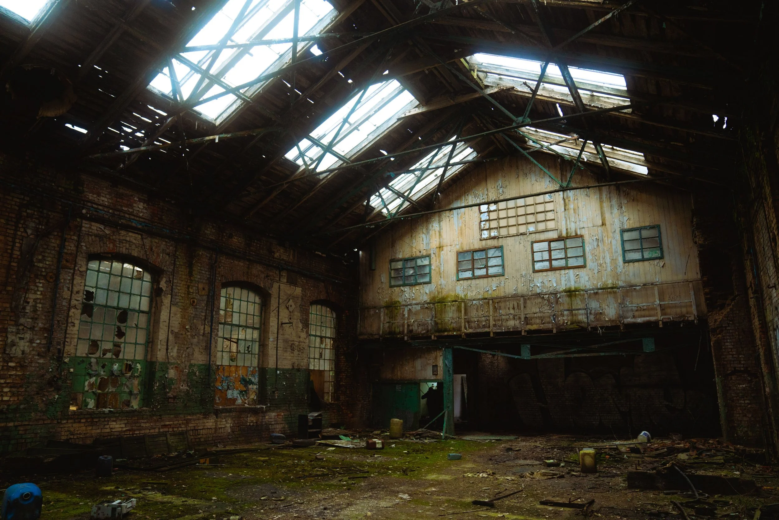An abandoned industrial building with a partially collapsed roof, broken windows, and moss-covered debris on the floor.