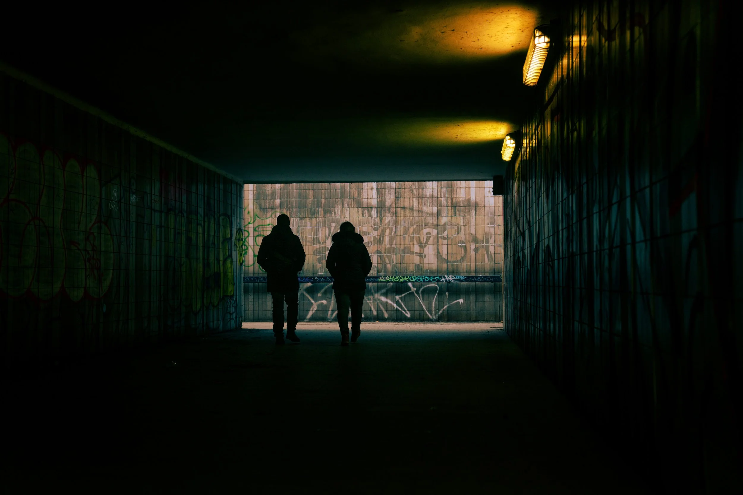 Two people walking through a dark, graffiti-covered underpass with dim lighting and a faint light at the far end.