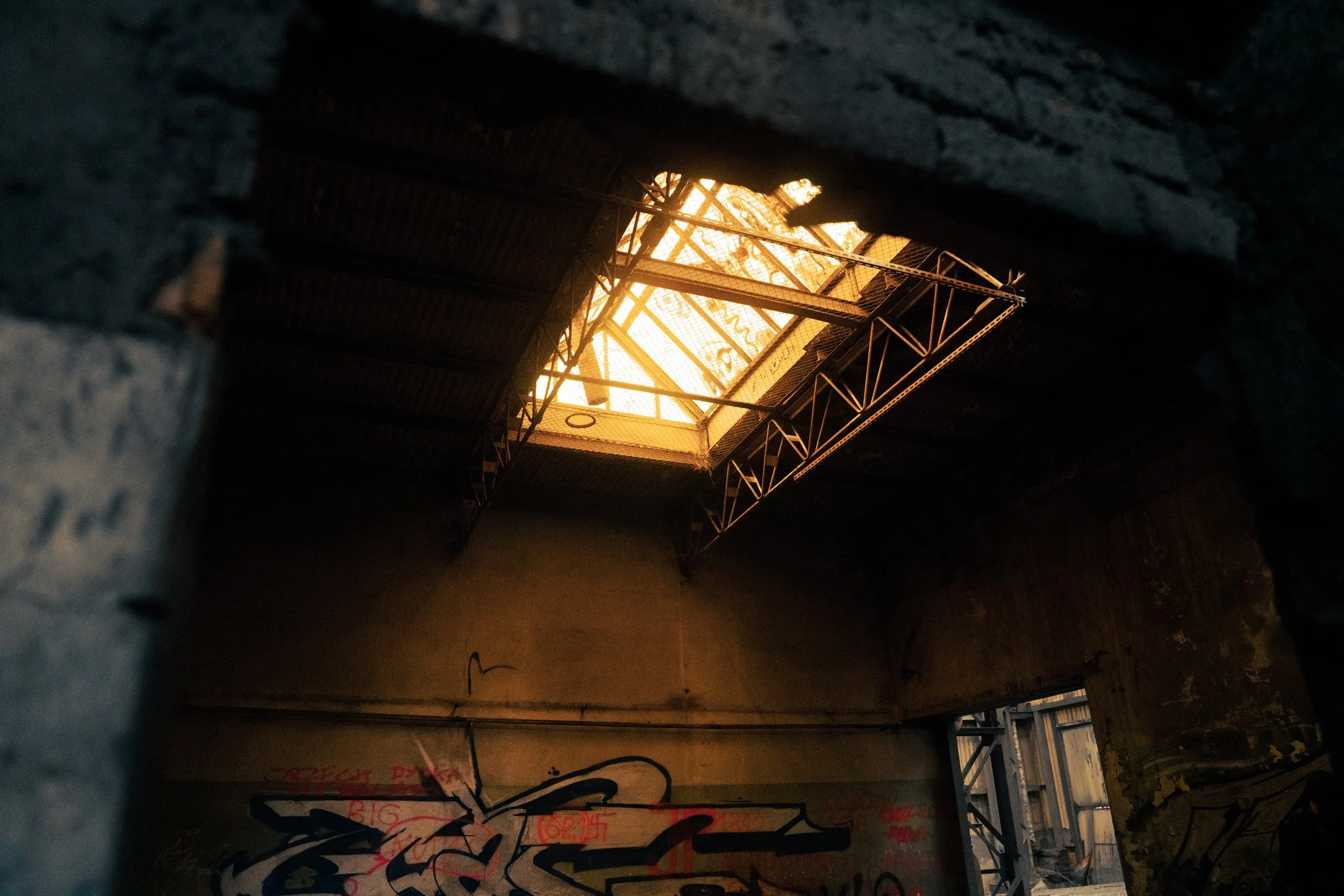 Looking up through a hole in the roof of an urban building with graffiti on the walls, revealing a bright, warm light from outside.