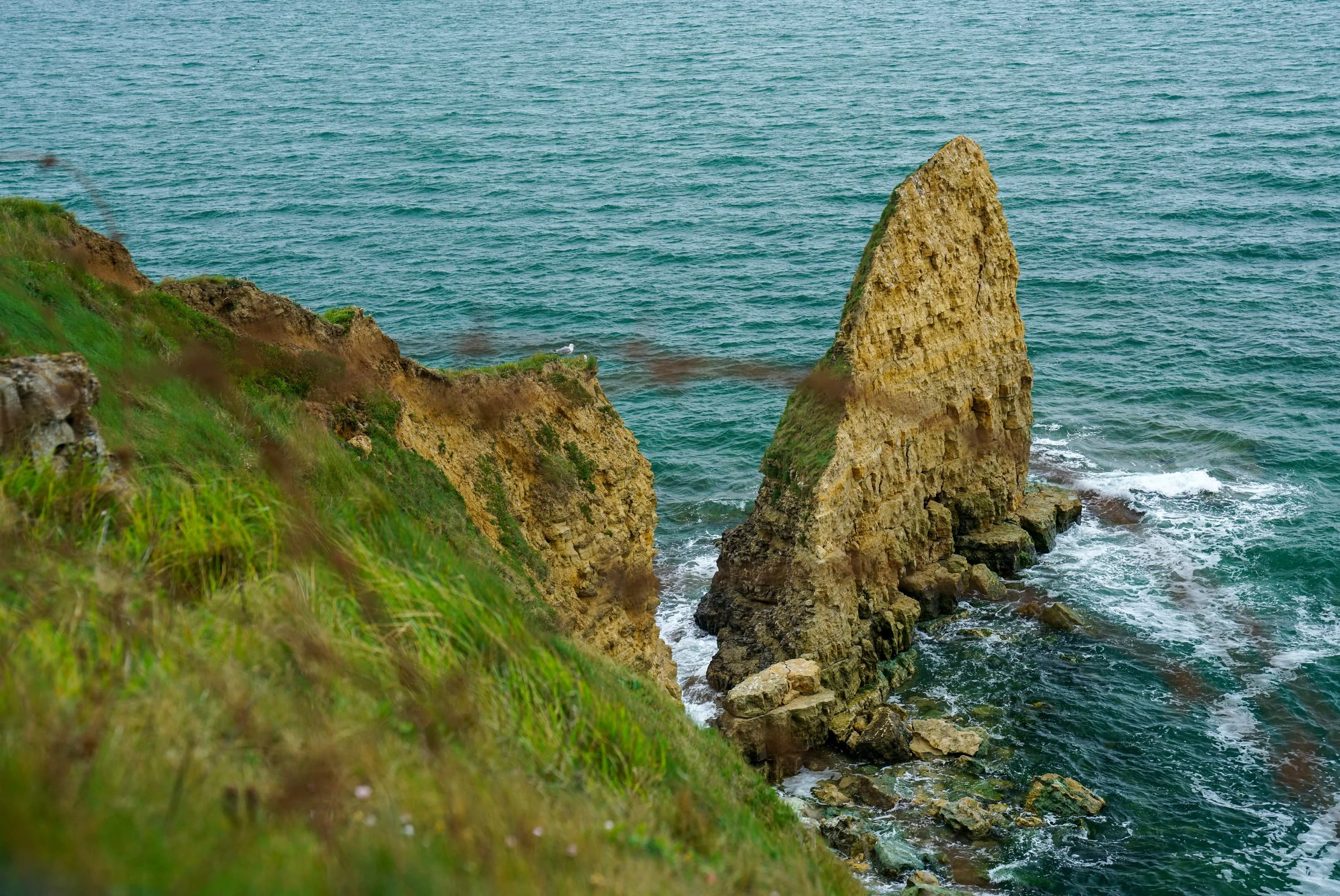 Cliffs with a large rock formation in the ocean surrounded by waves and green grass on the cliff edge.