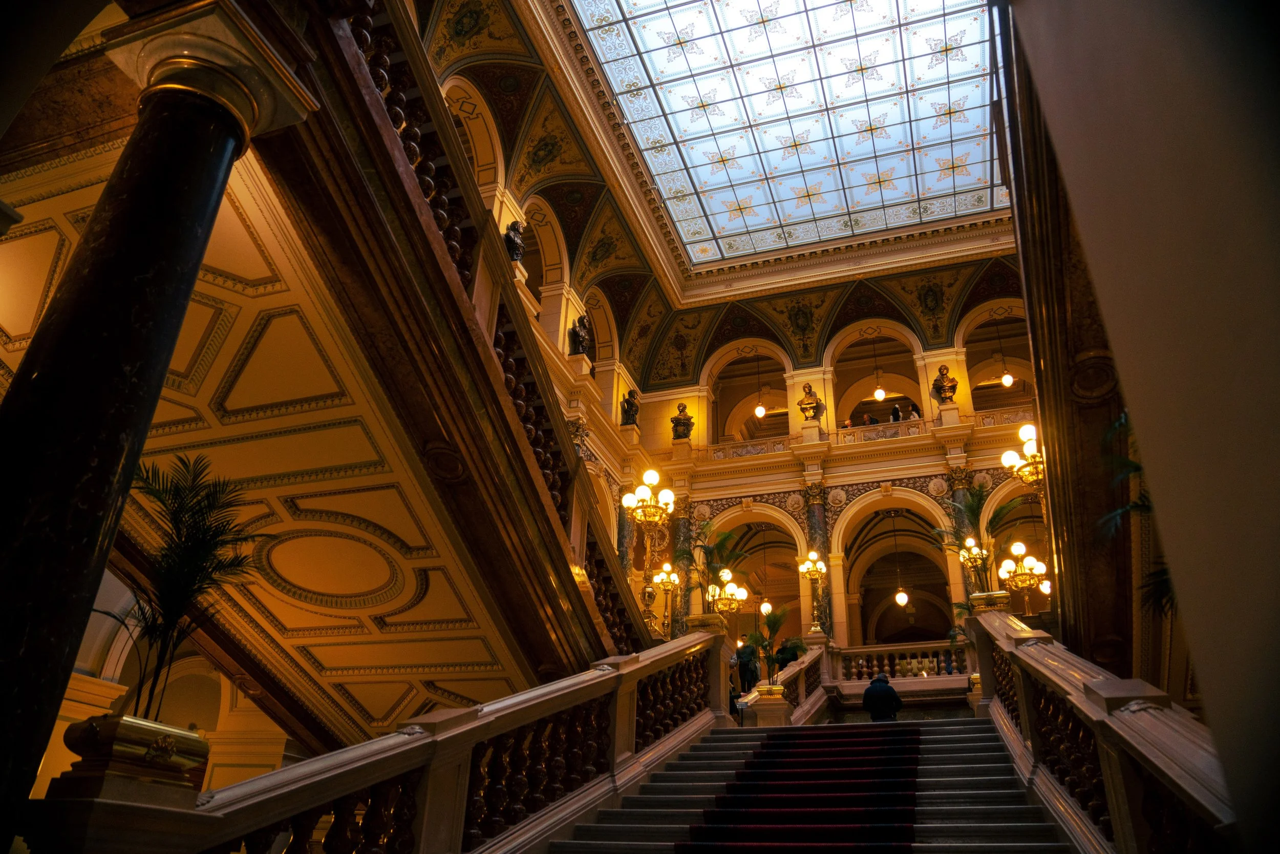 Interior view of a grand, historic building with a staircase leading upwards, ornate ceiling with decorative patterns, large glass skylight, chandeliers, and classical statues along the upper balconies.