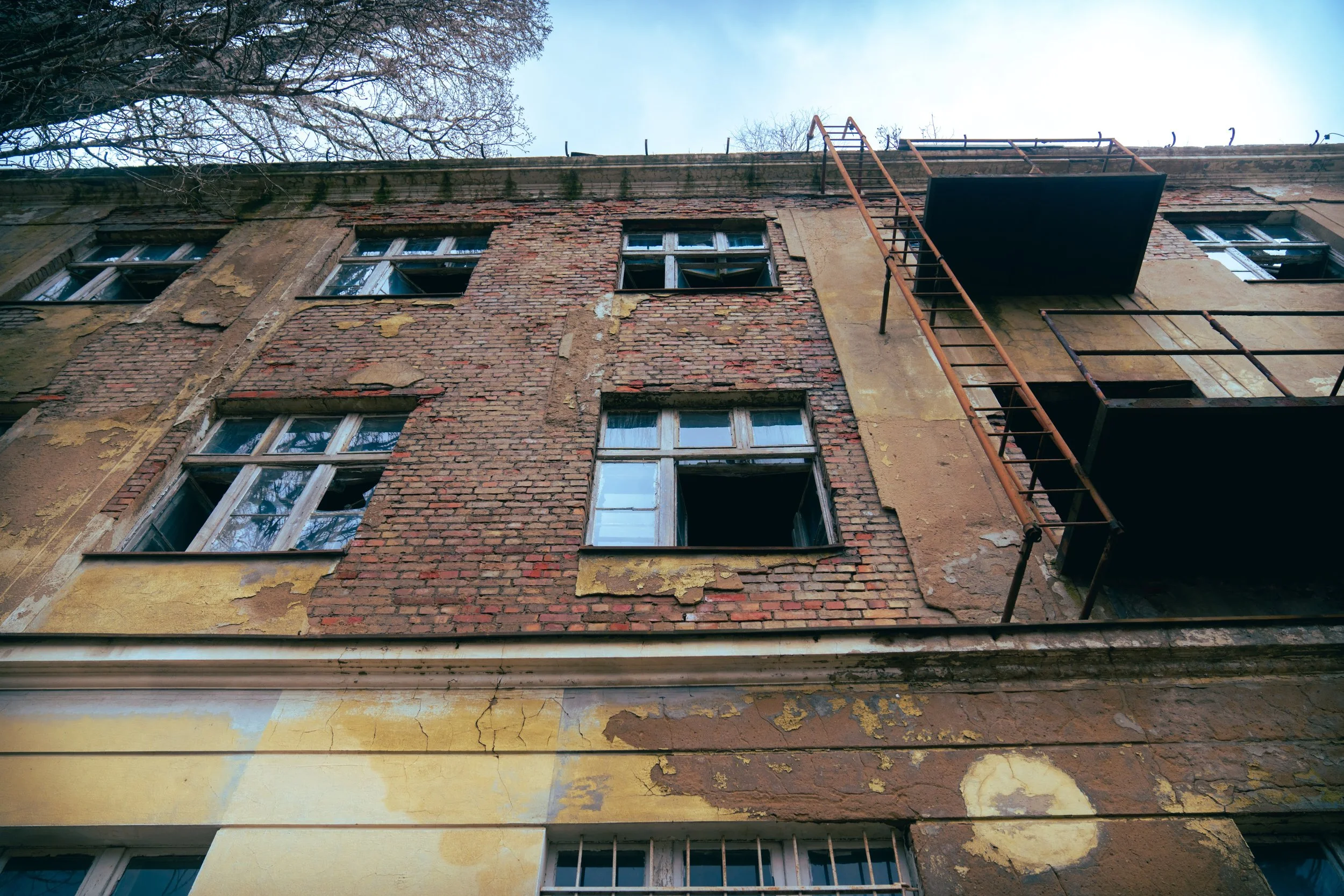 The exterior of an old building with peeling yellow and brick walls, some broken windows, and rusted fire escapes.